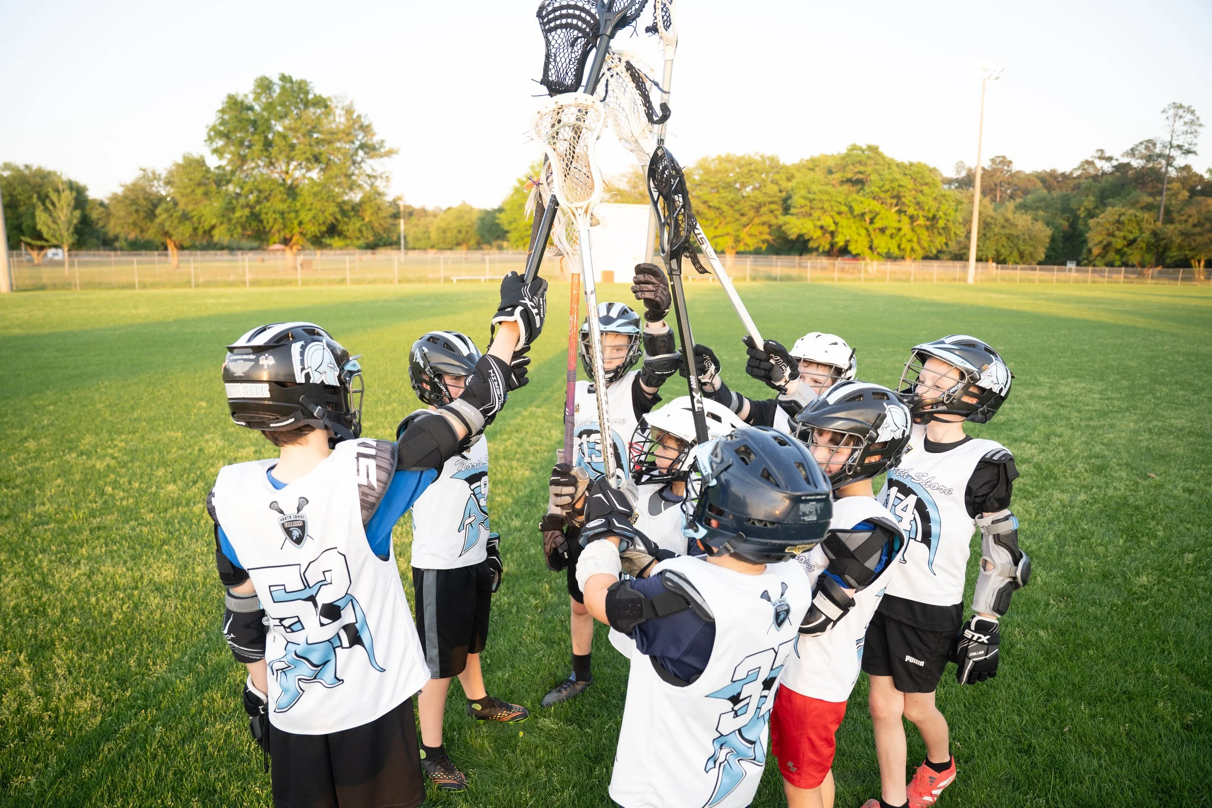 A group of young boys in hockey gear, including helmets and jerseys, raising their hockey sticks together in a huddle on a grassy field during daytime with trees and a fence in the background.