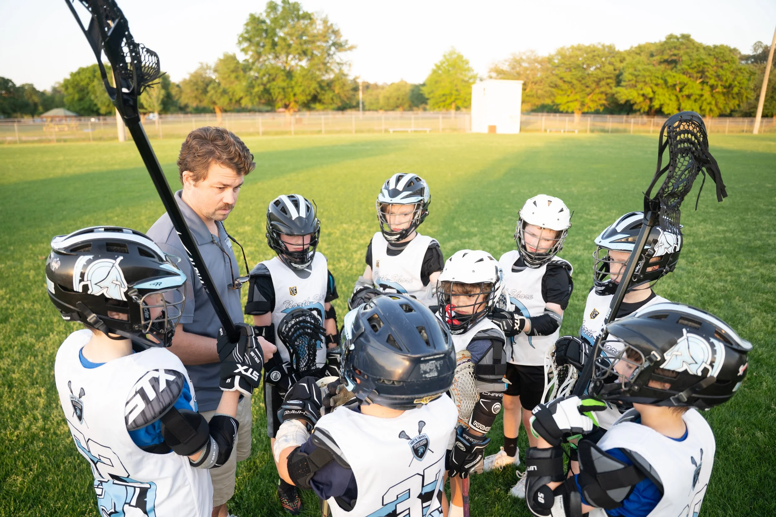 A youth hockey coach giving instructions to a group of young children huddled together on a grassy field, all wearing hockey gear and helmets.