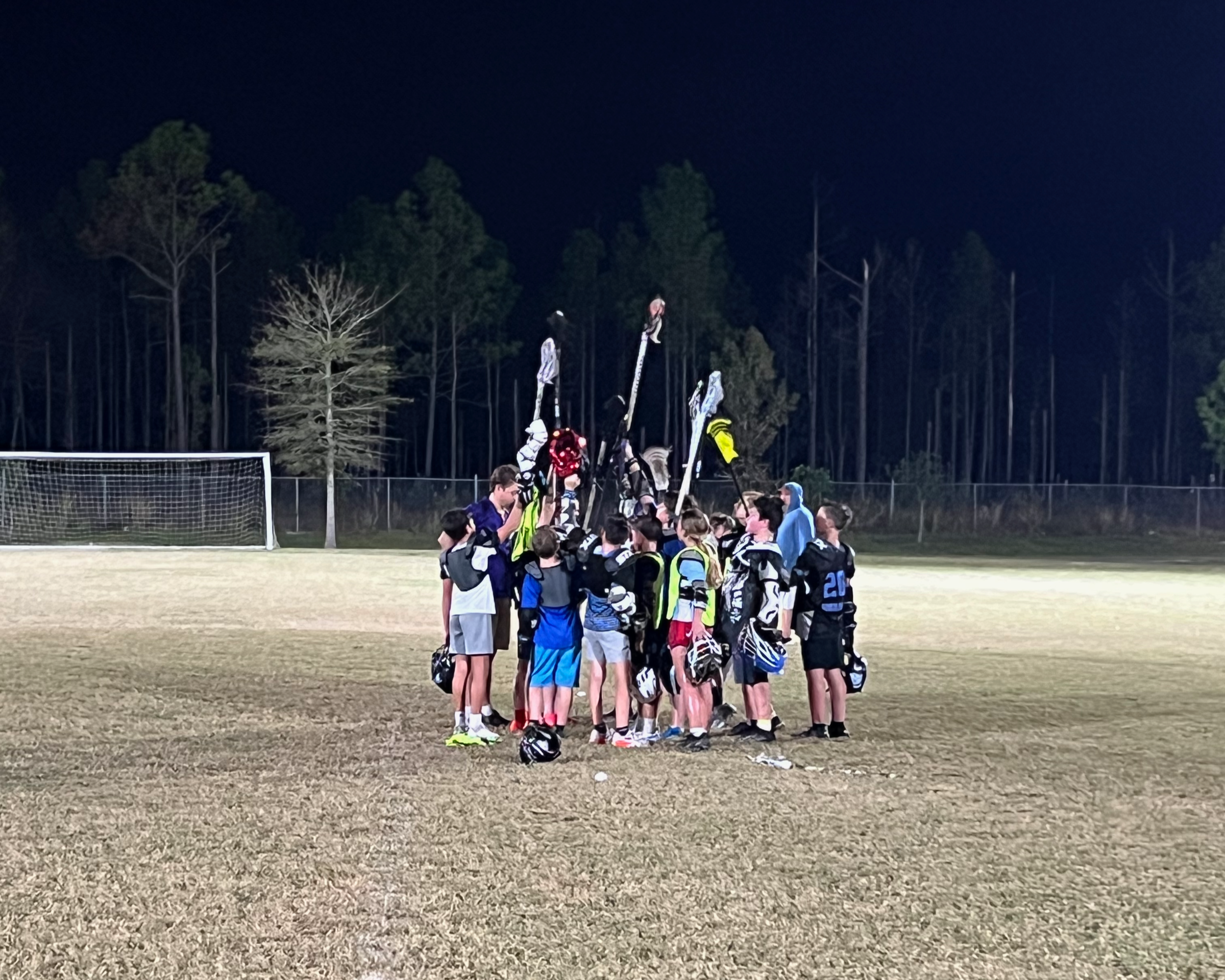 A group of youth athletes in sports uniforms and backpacks forming a human pyramid on a sports field at night, with a goalpost and trees in the background.