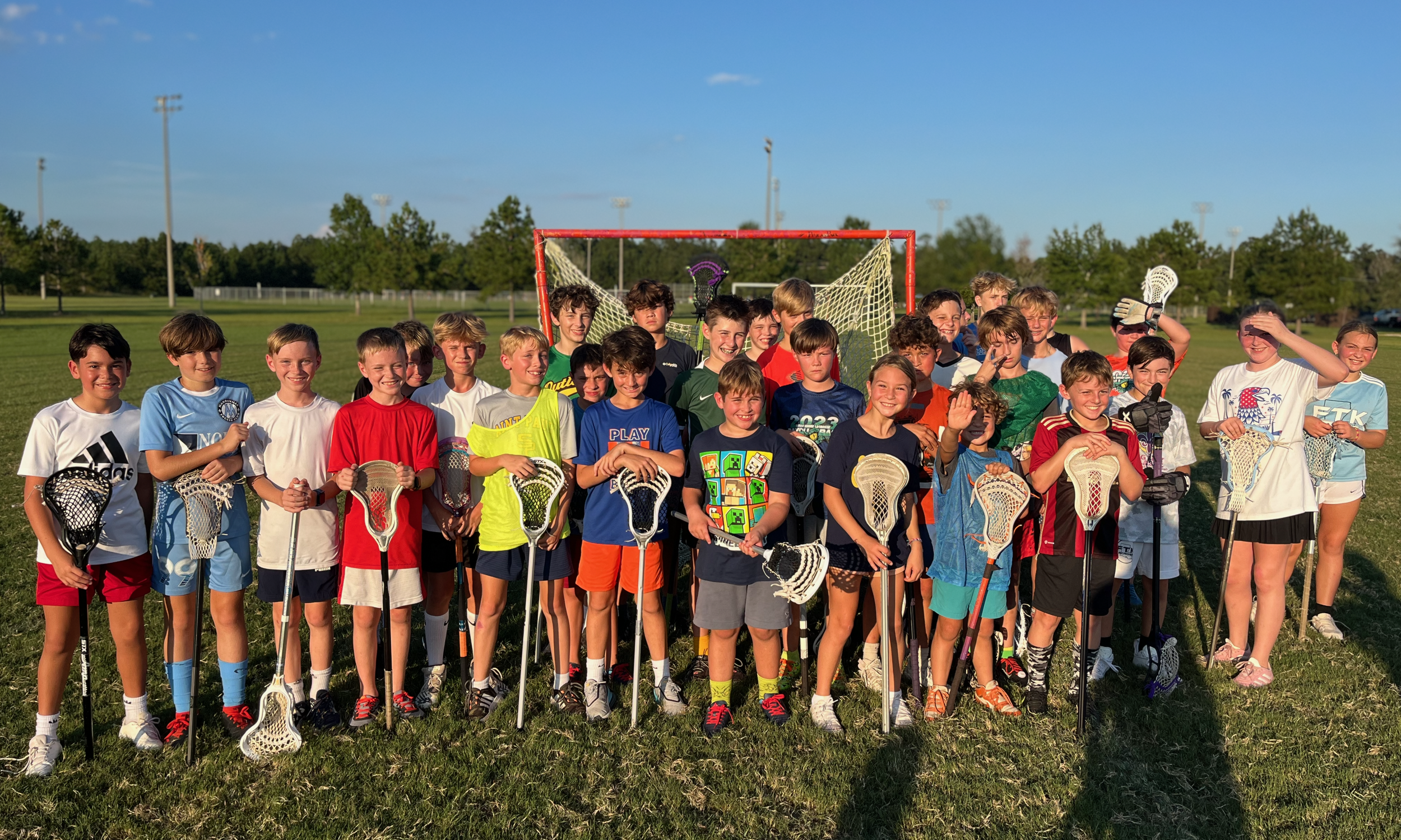 Group of children in sports uniforms holding lacrosse sticks on a grassy field with a goalpost in the background during daytime.
