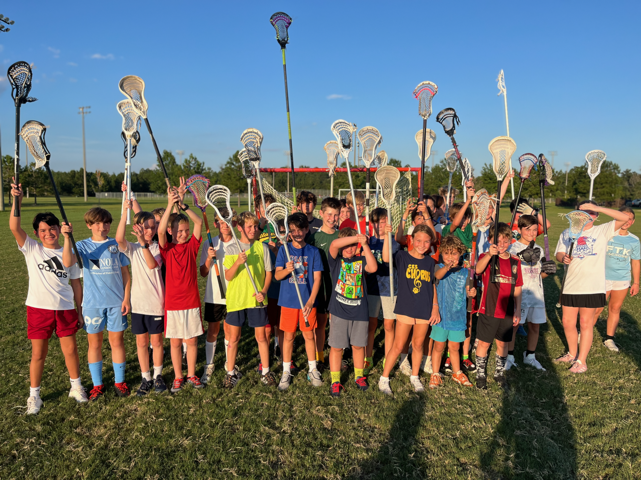 Group of young boys and girls holding lacrosse sticks on a grassy field, smiling, with a clear blue sky above.