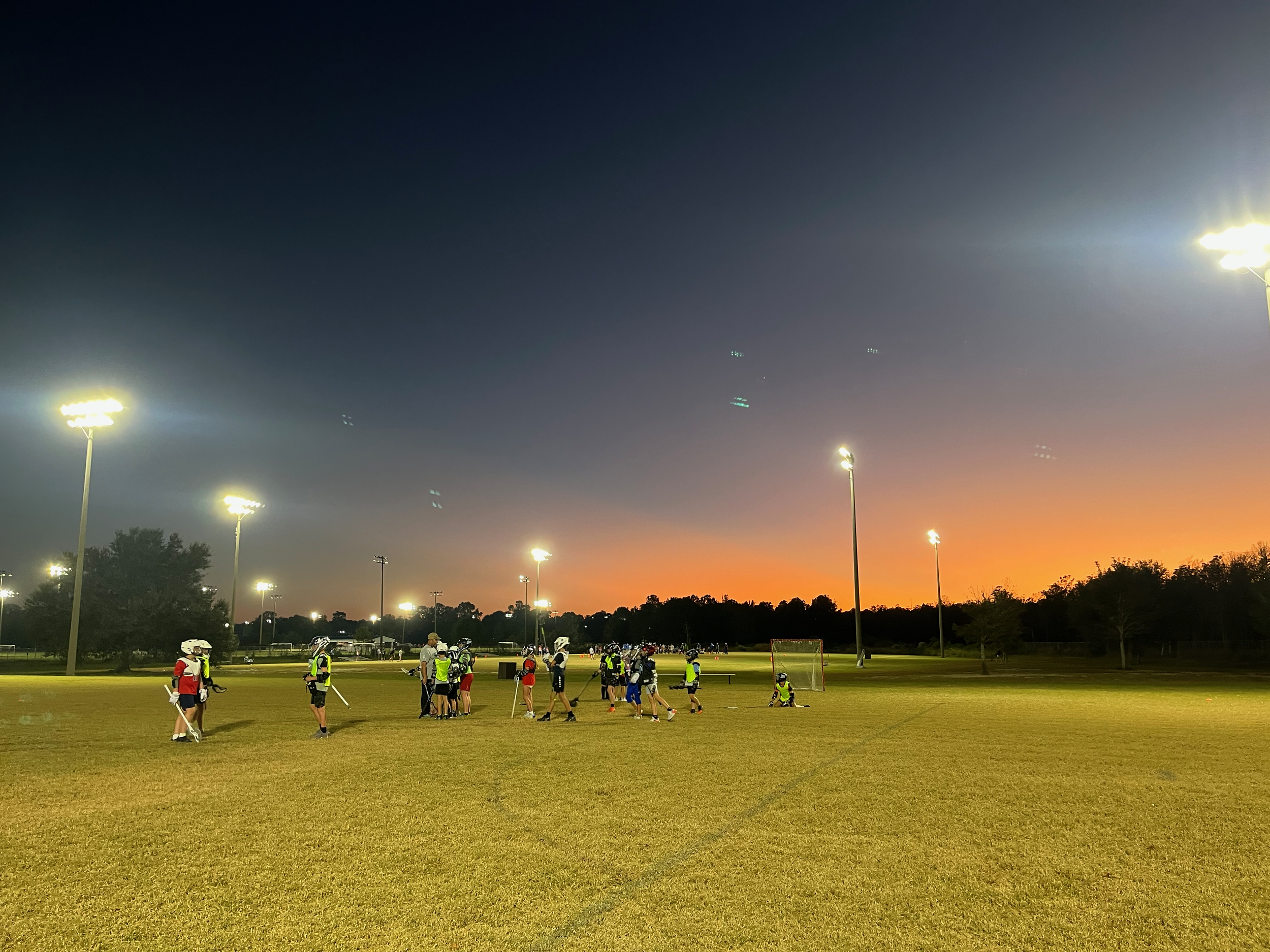 A dusk scene of a sports field with bright floodlights illuminating the area, where young athletes in protective gear are practicing or playing. The background features a darkening sky with a hint of sunset at the horizon, and silhouettes of trees.