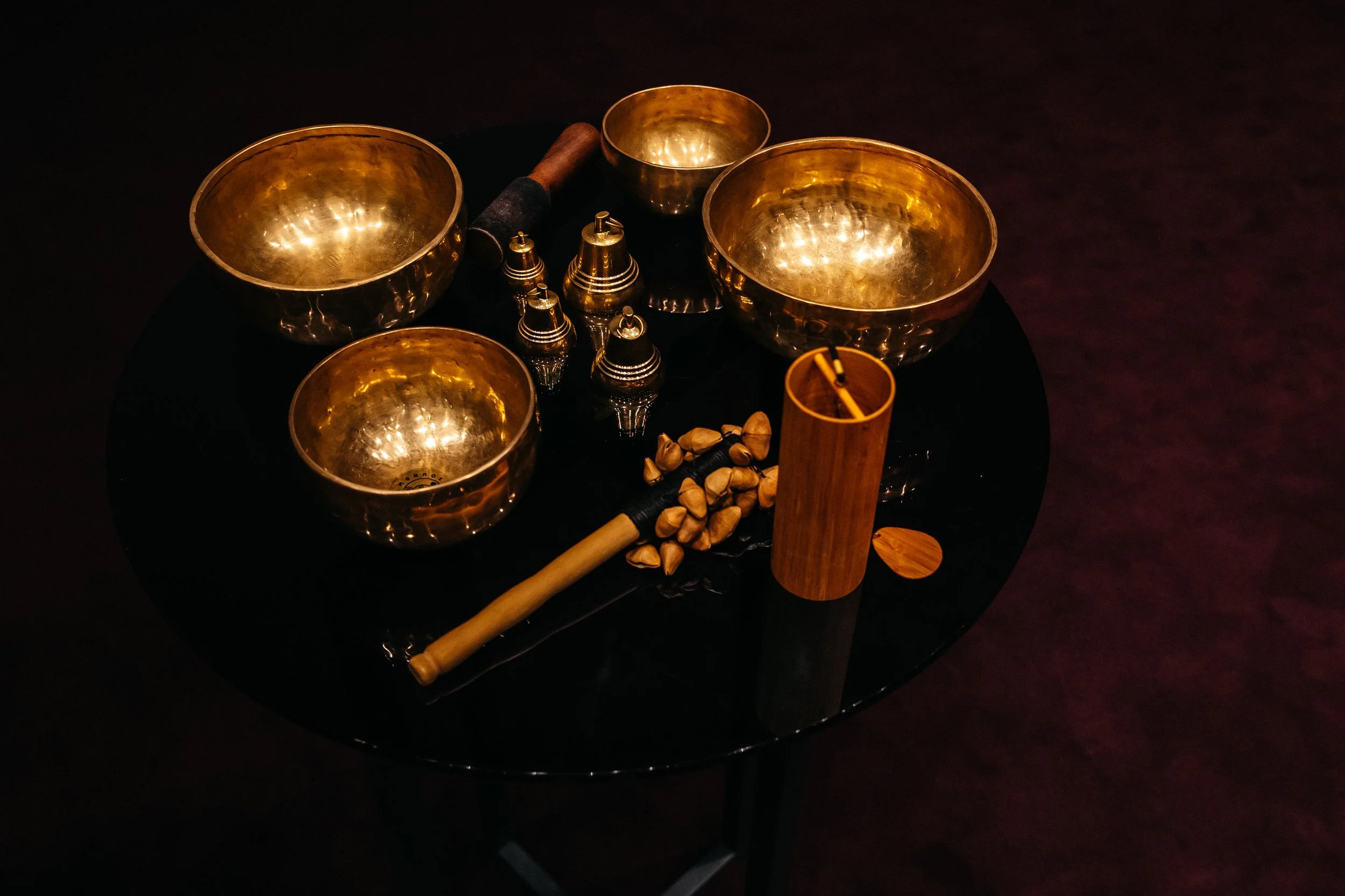 A black table with four gold singing bowls, a wooden stick, small metallic bells, a wooden cylinder, and a few small stones.