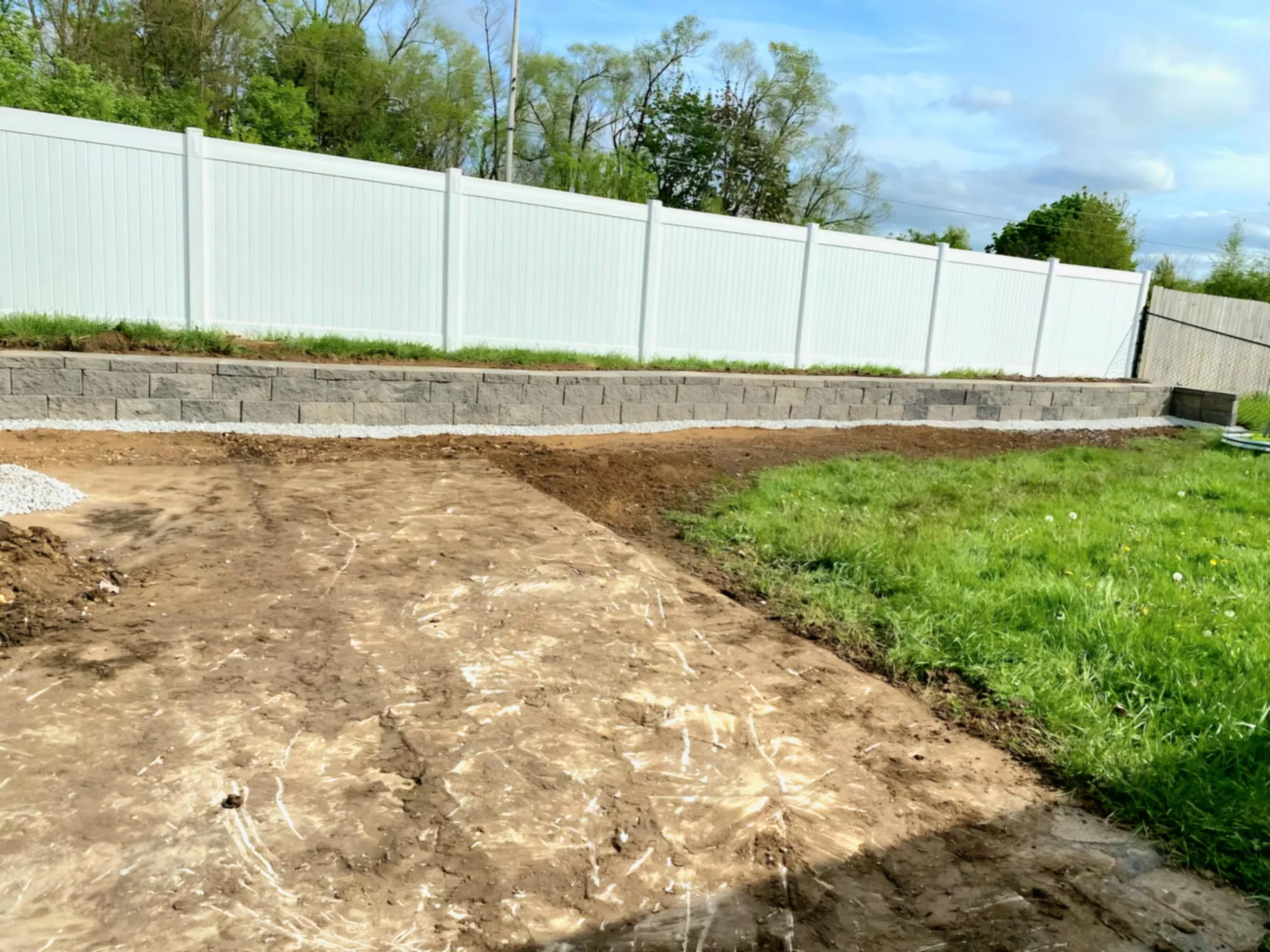 A backyard with a dirt path or patio, a retaining wall made of concrete blocks, a white fence, green grass, and trees in the background under a partly cloudy sky.