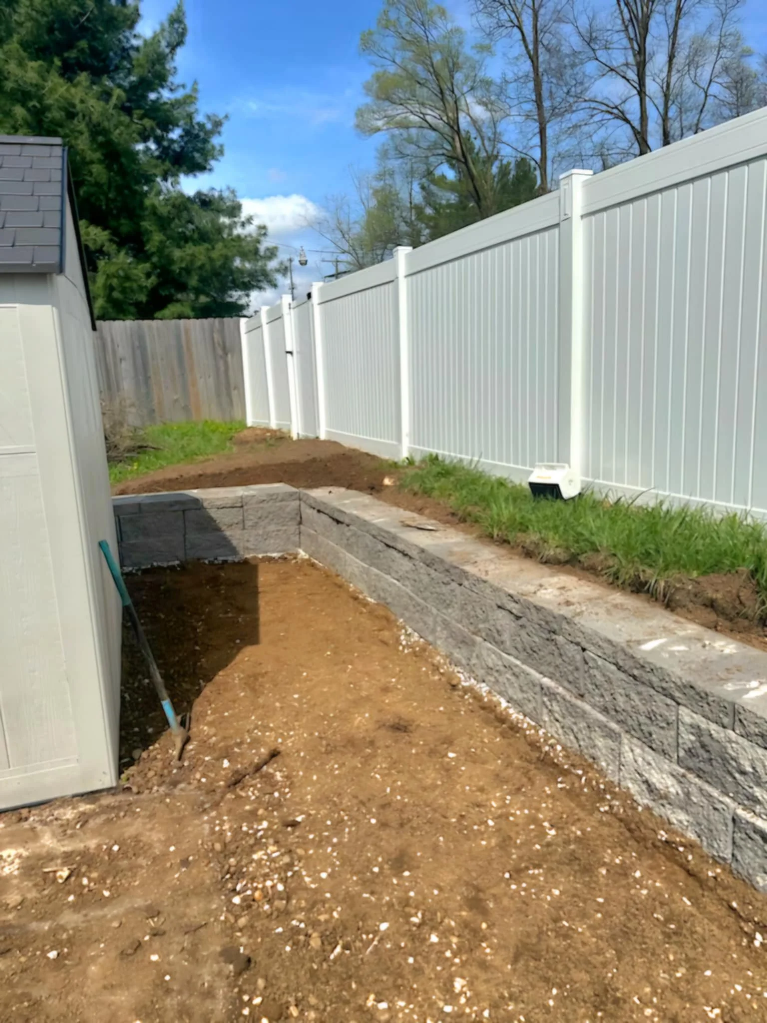 Backyard with a white vinyl privacy fence, garden bed with stone retaining wall, and a small shed on the left, under a partly cloudy sky.