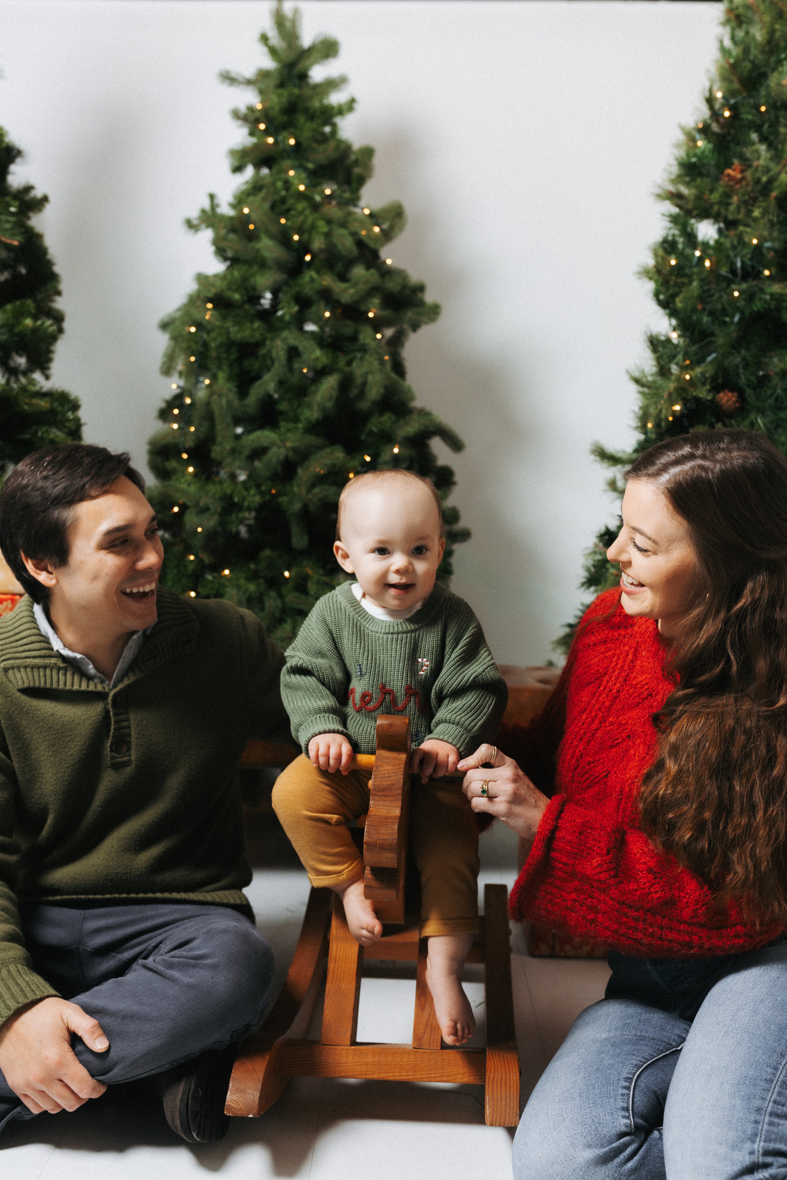 A family celebrating Christmas, with a baby sitting on a wooden rocking horse, surrounded by decorated Christmas trees and smiling adults.