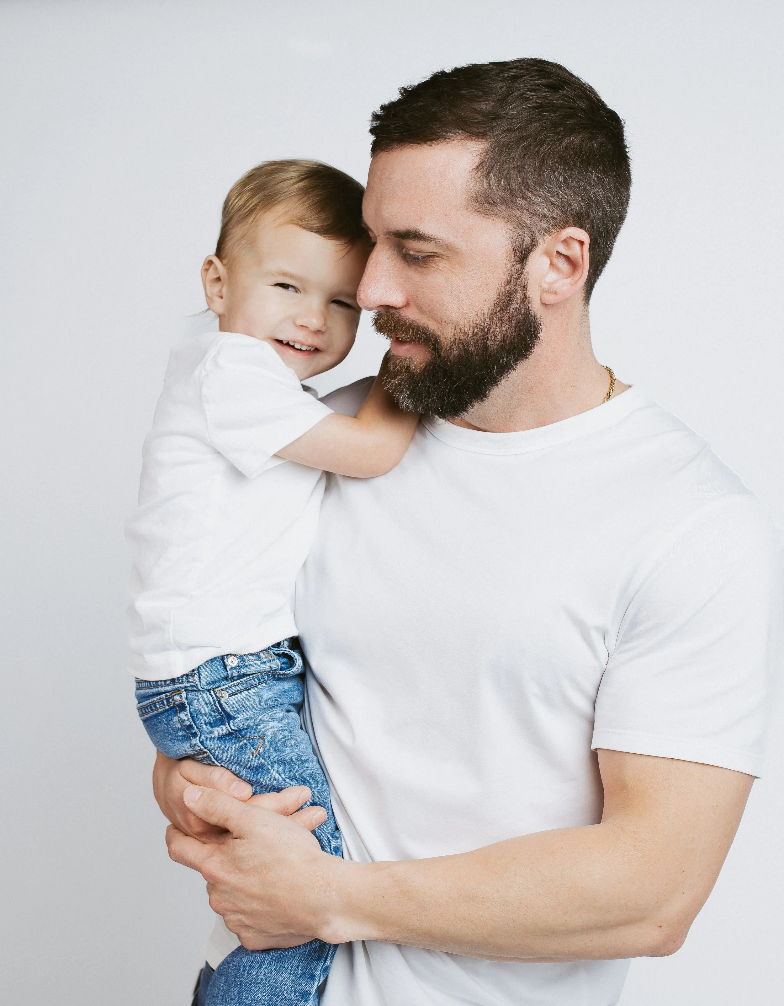 A man with a beard holding a smiling young boy with blonde hair, both wearing white shirts against a plain background.