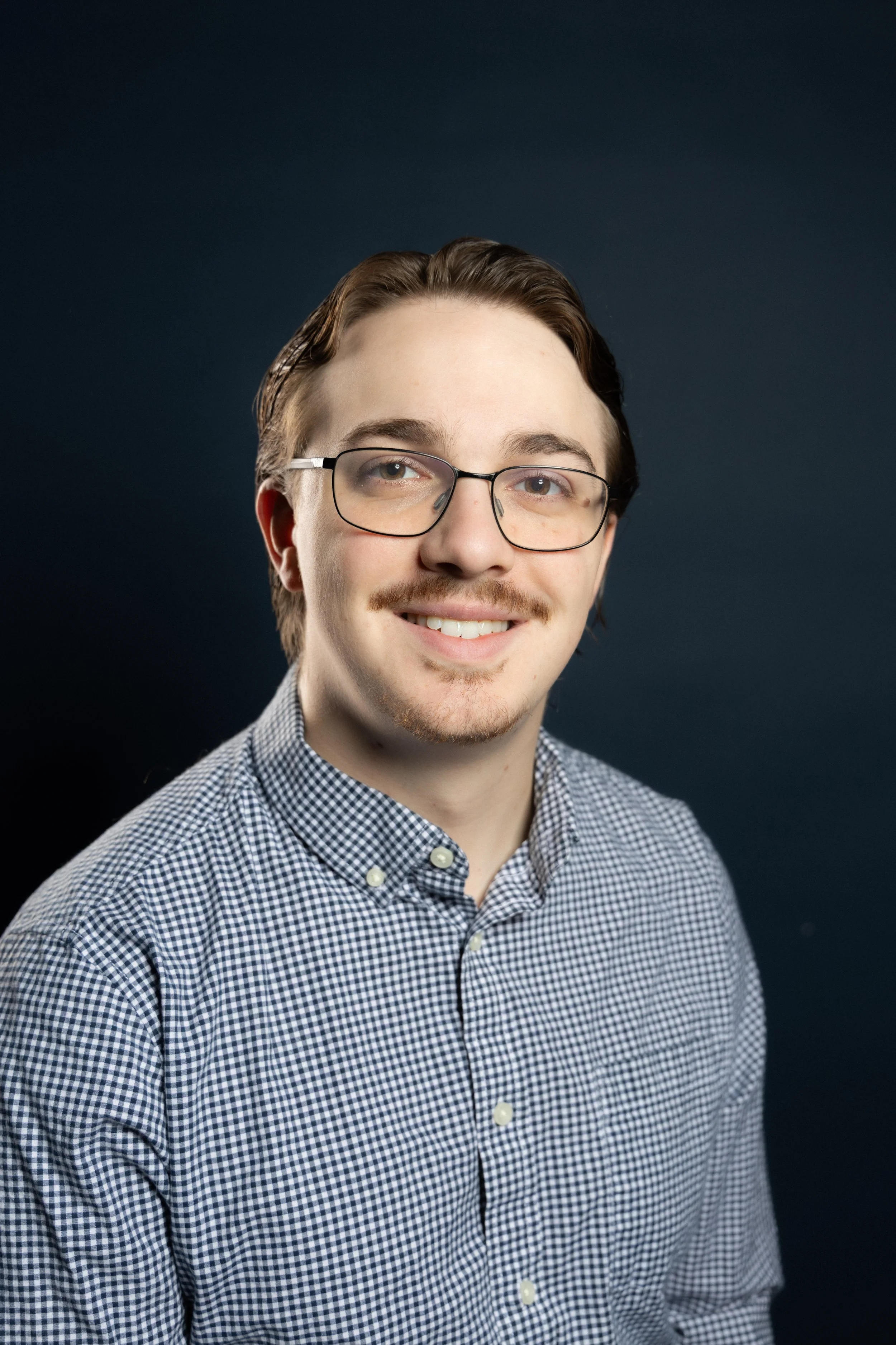 A young man with glasses, a mustache, and a goatee, smiling, wearing a checkered button-up shirt, against a dark background.