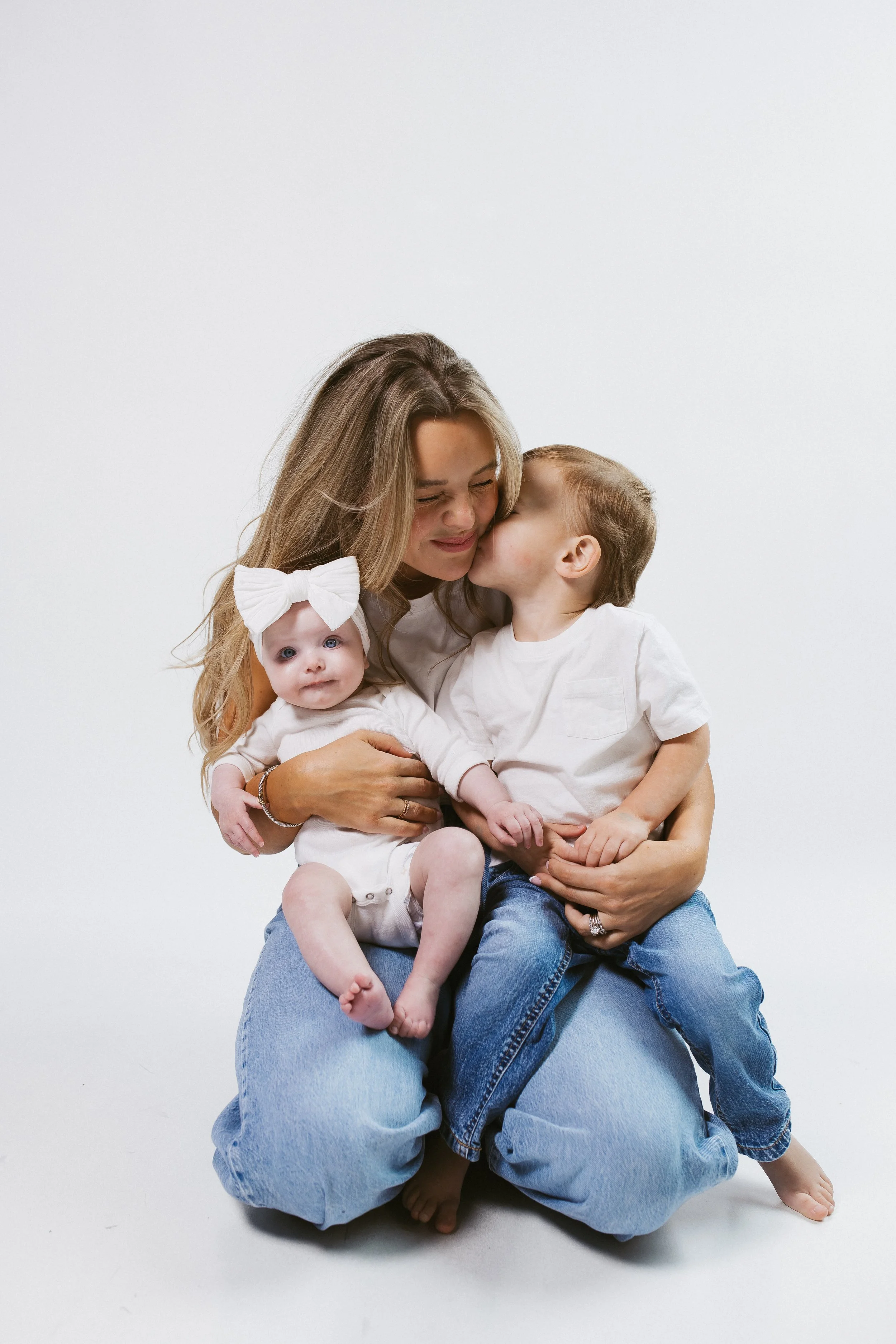 A woman with long, wavy blonde hair holds two children, a young girl with a large white bow on her head and a boy, both wearing white shirts and blue jeans, against a plain white background.