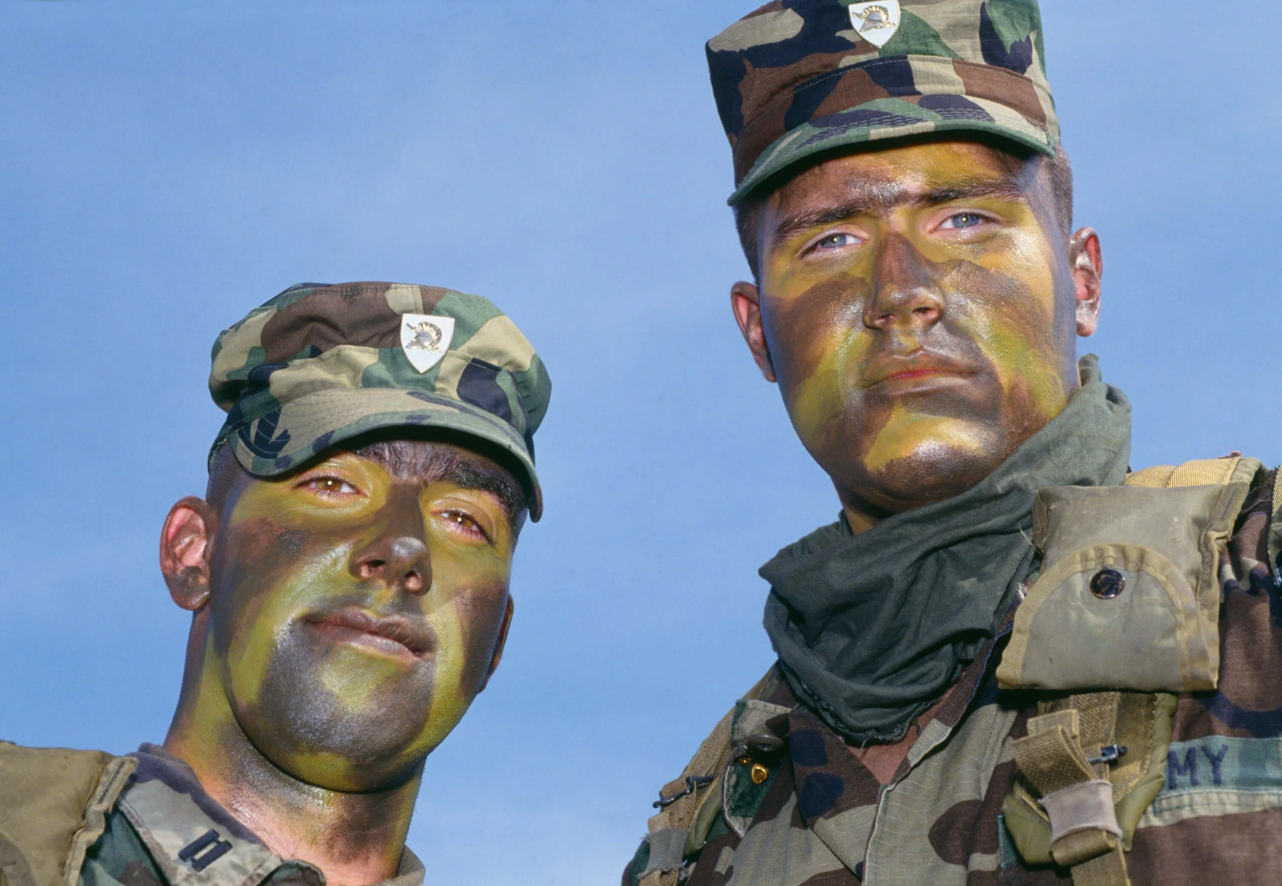 Two soldiers in camouflage uniforms and hats with face paint standing outdoors against a blue sky.