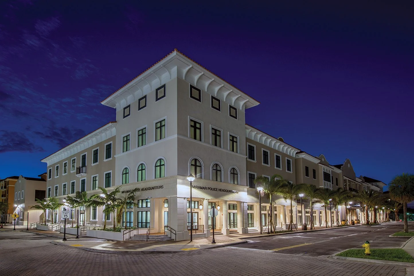 Night view of the Miramar Police Headquarters building with palm trees lining the sidewalk, illuminated street lamps, and a clear evening sky.