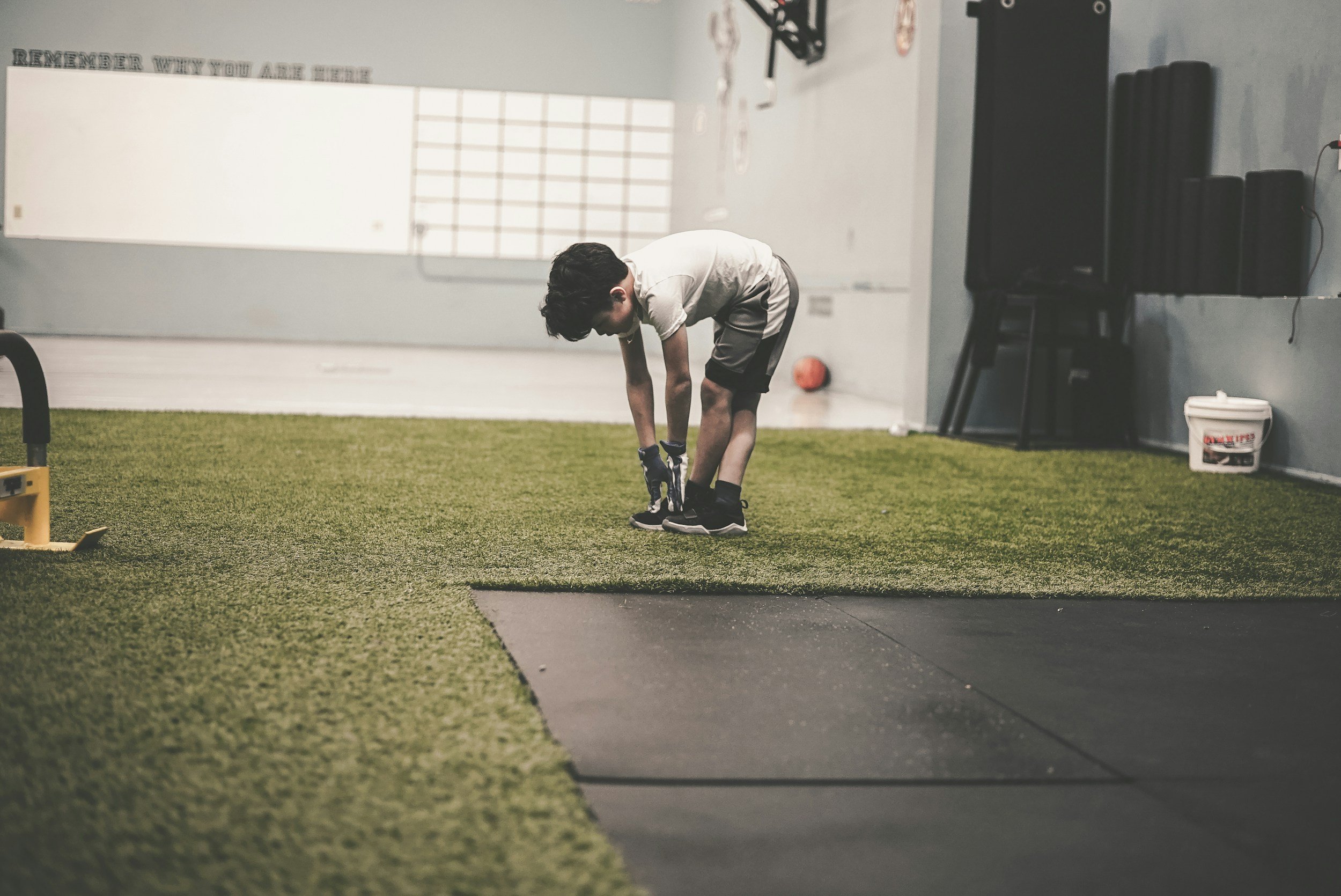 A young boy in a gym, bending over to tie his shoelaces on a green artificial turf floor.