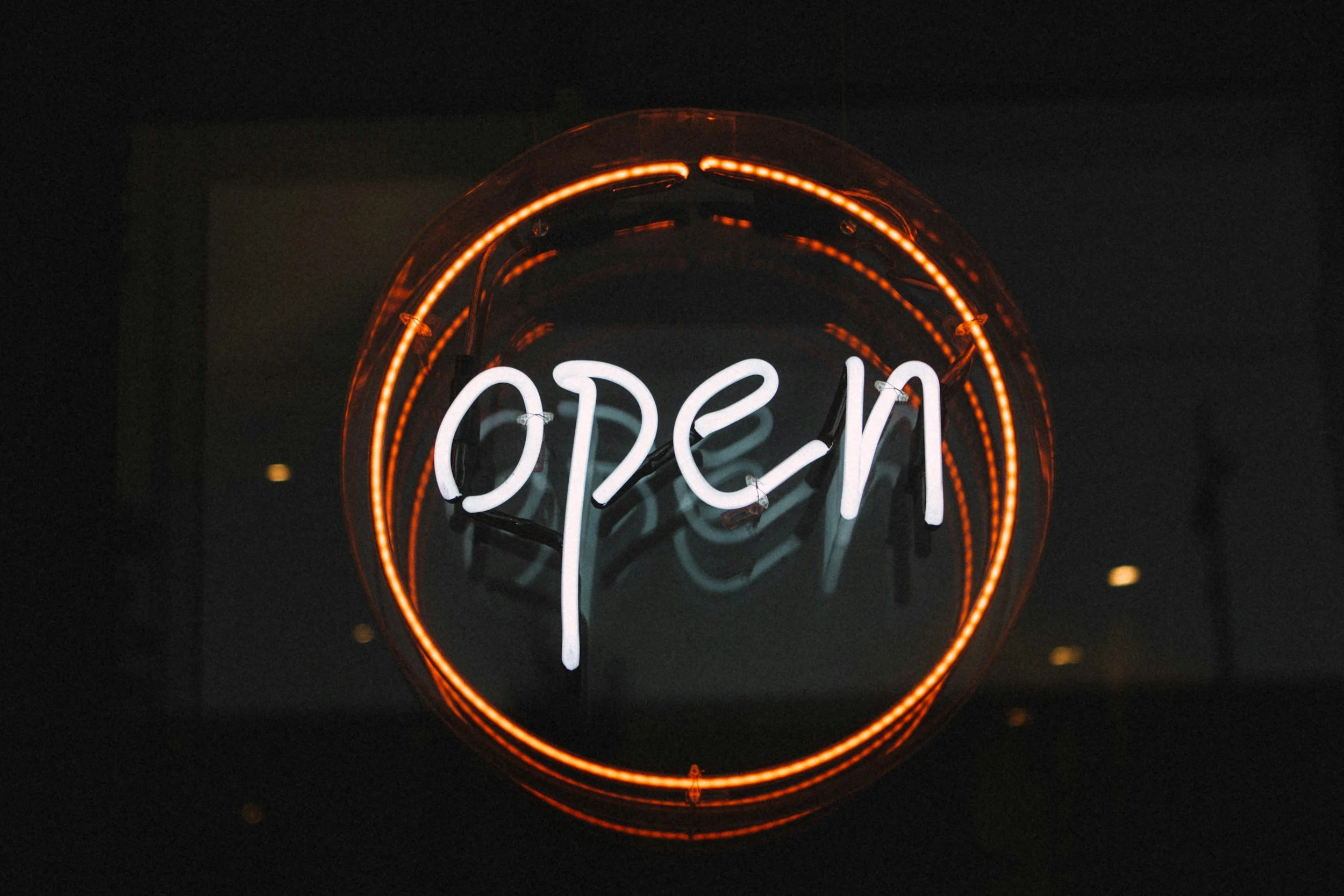 Backlit neon sign displaying the word 'OPEN' in white neon lights, surrounded by a circular orange neon light.