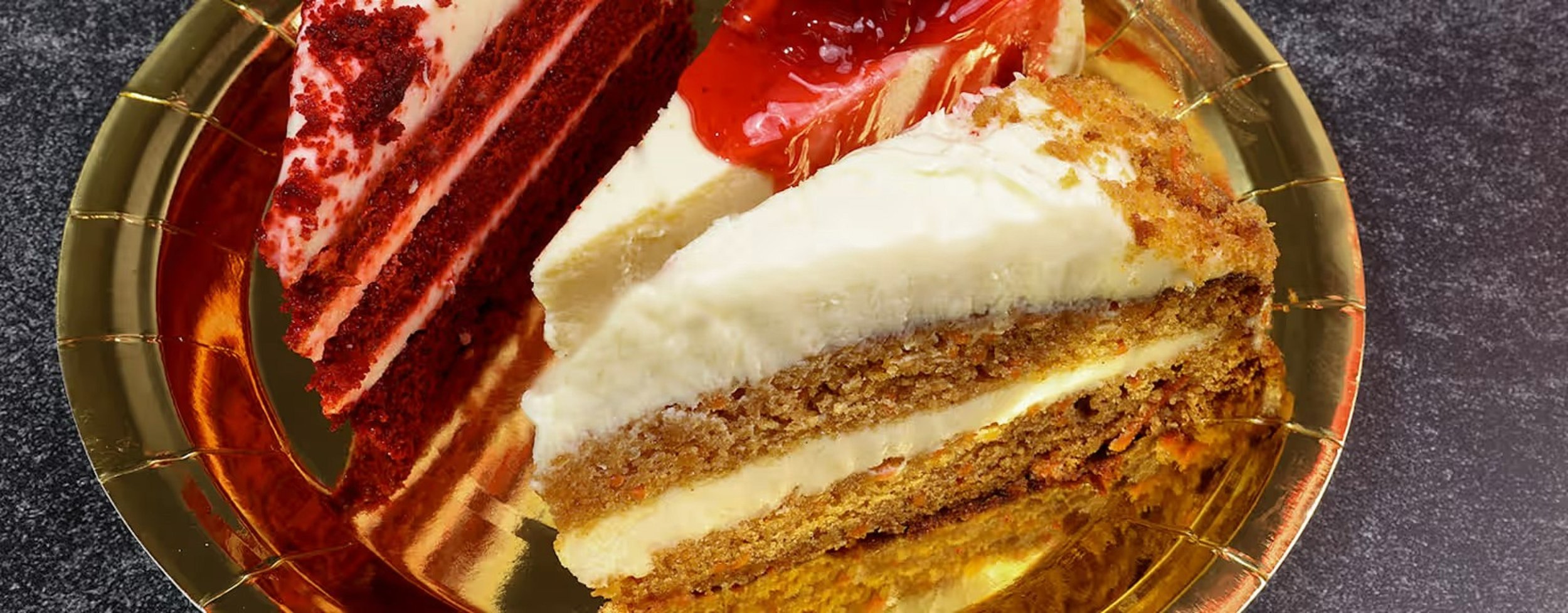Three slices of different layered cakes on a golden plate, with a dark table surface underneath.