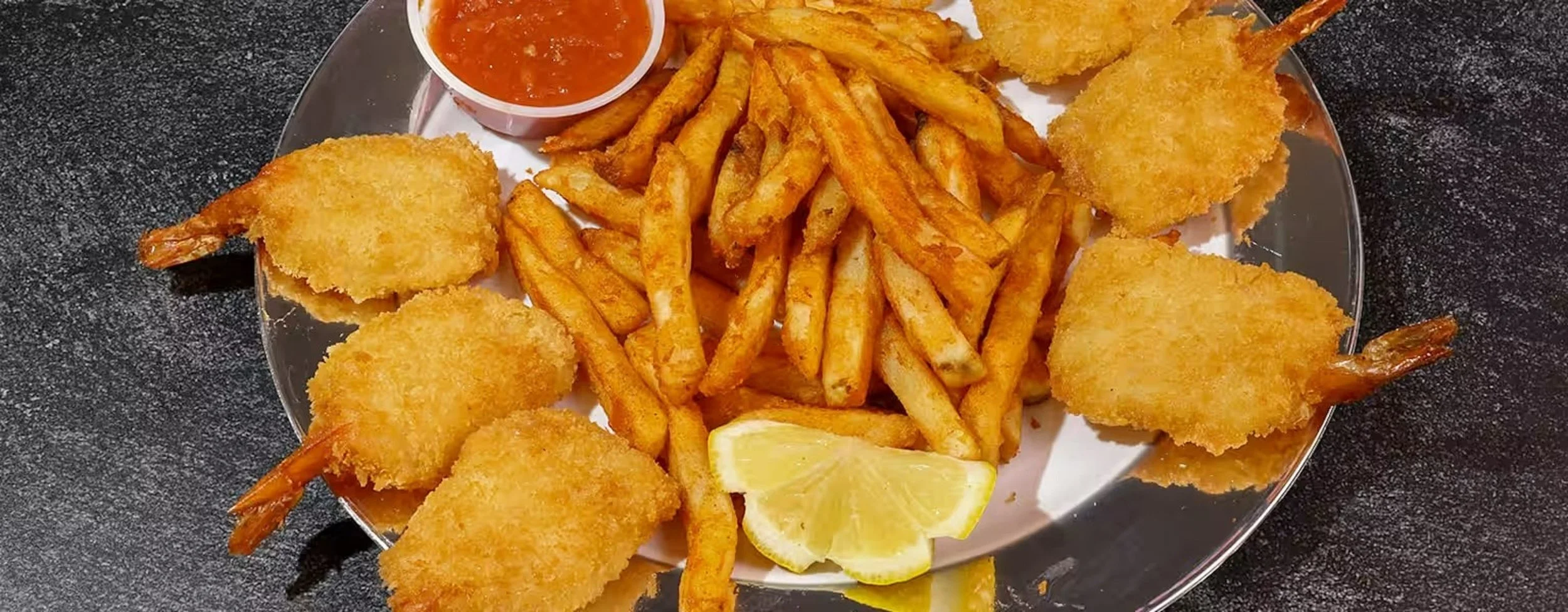 Plate of fried shrimp, French fries, lemon wedges, and a small cup of cocktail sauce on a black surface.