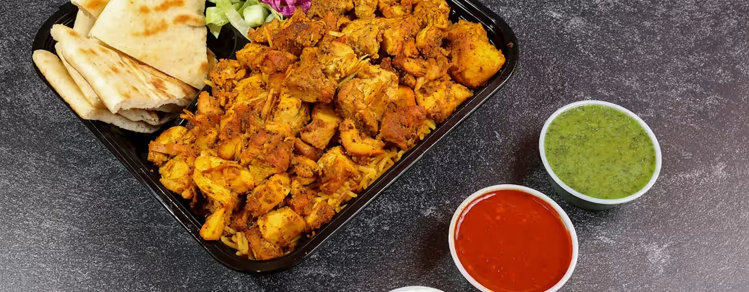 A meal served in a black tray including spicy chicken, naan bread, and a small salad with two cups of green chutney and red sauce on a black surface.