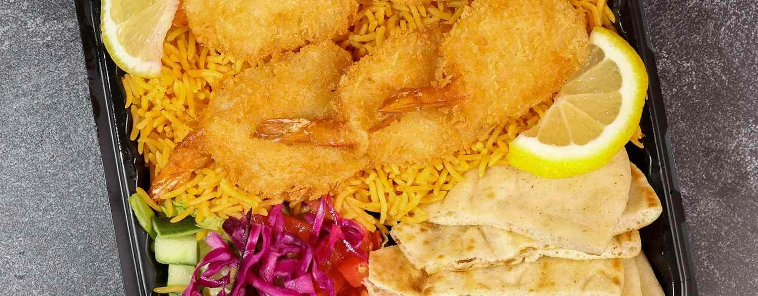 A takeout tray with breaded shrimp, yellow rice, lemon wedges, sliced flatbread, and side salad with cucumbers and purple cabbage.