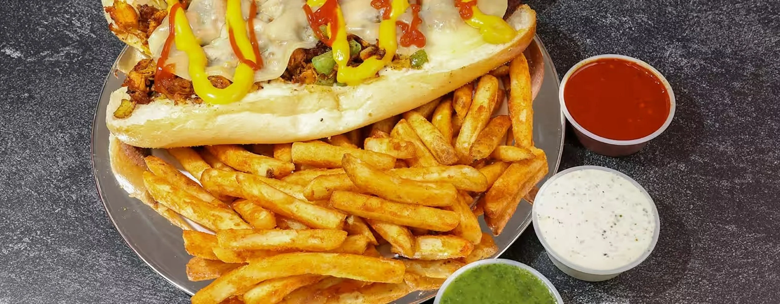 Philly cheesesteak sandwich with French fries, served with three dipping sauces on a metal plate.
