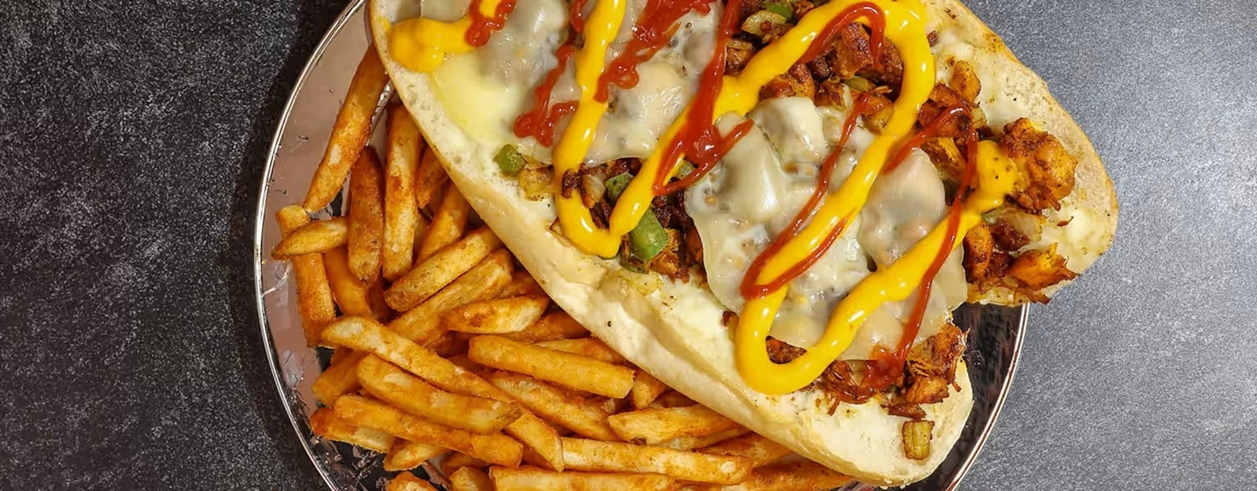 Philly cheesesteak sandwich topped with yellow and red sauces, served with French fries on a silver platter.