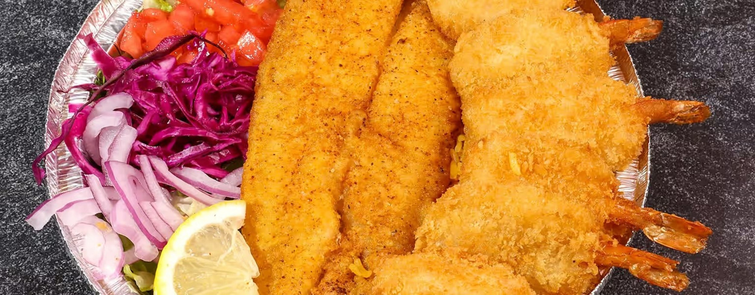 Plate with breaded fried fish, two pieces of fried shrimp, sliced lemon, purple cabbage slaw, tomato slices, and sliced onions on a black countertop.