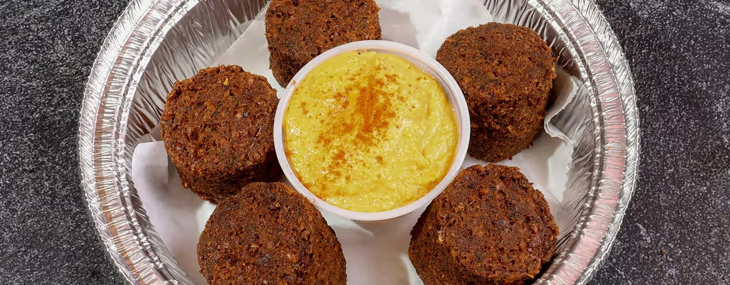 Fried falafel balls arranged on a paper-lined foil tray around a small container of yellow dipping sauce with a sprinkle of spice on top, on a dark speckled countertop.