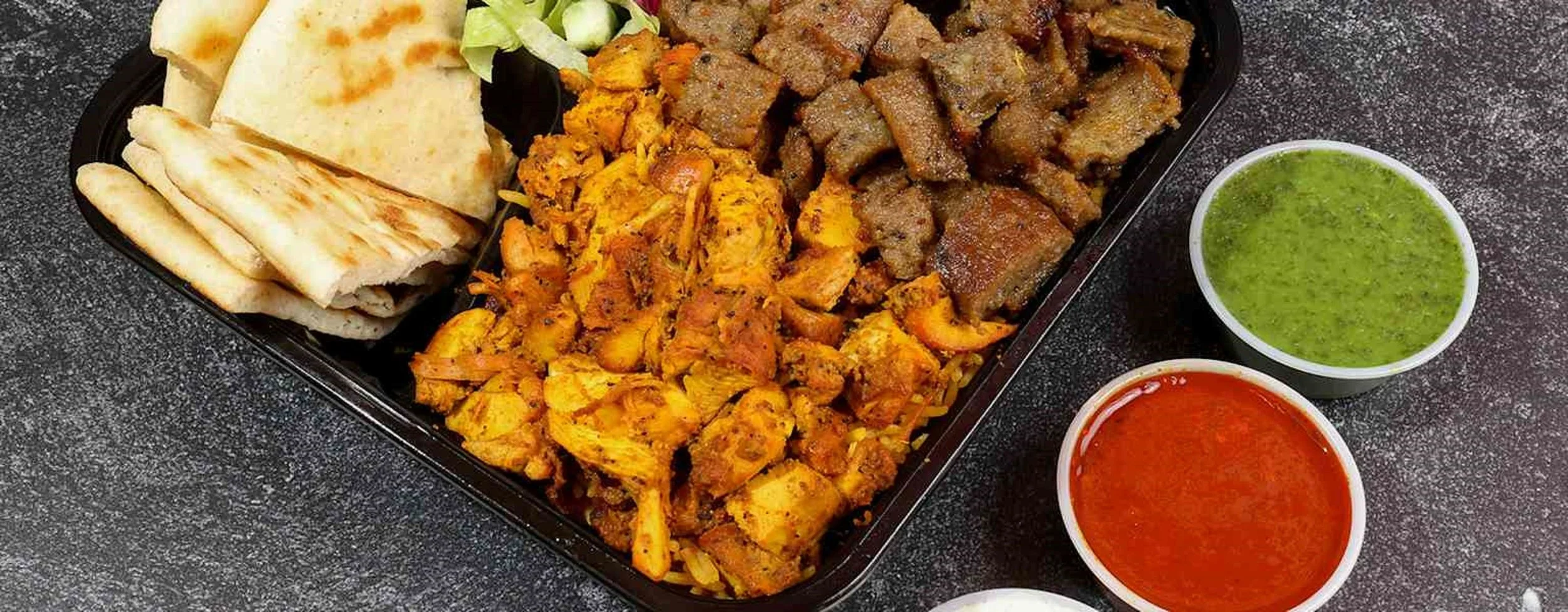 Cuisine platter with pita bread, salad, and four types of cooked meats with two bowls of green and red sauces.