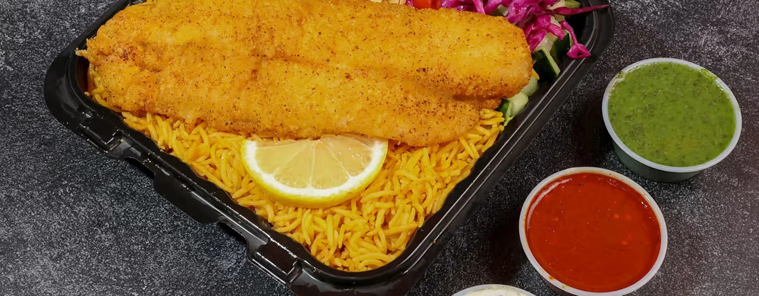A black tray with seasoned yellow rice topped with a large breaded fish fillet, a lemon wedge, and colorful vegetables. Two small cups of green and red sauces are placed next to the tray.