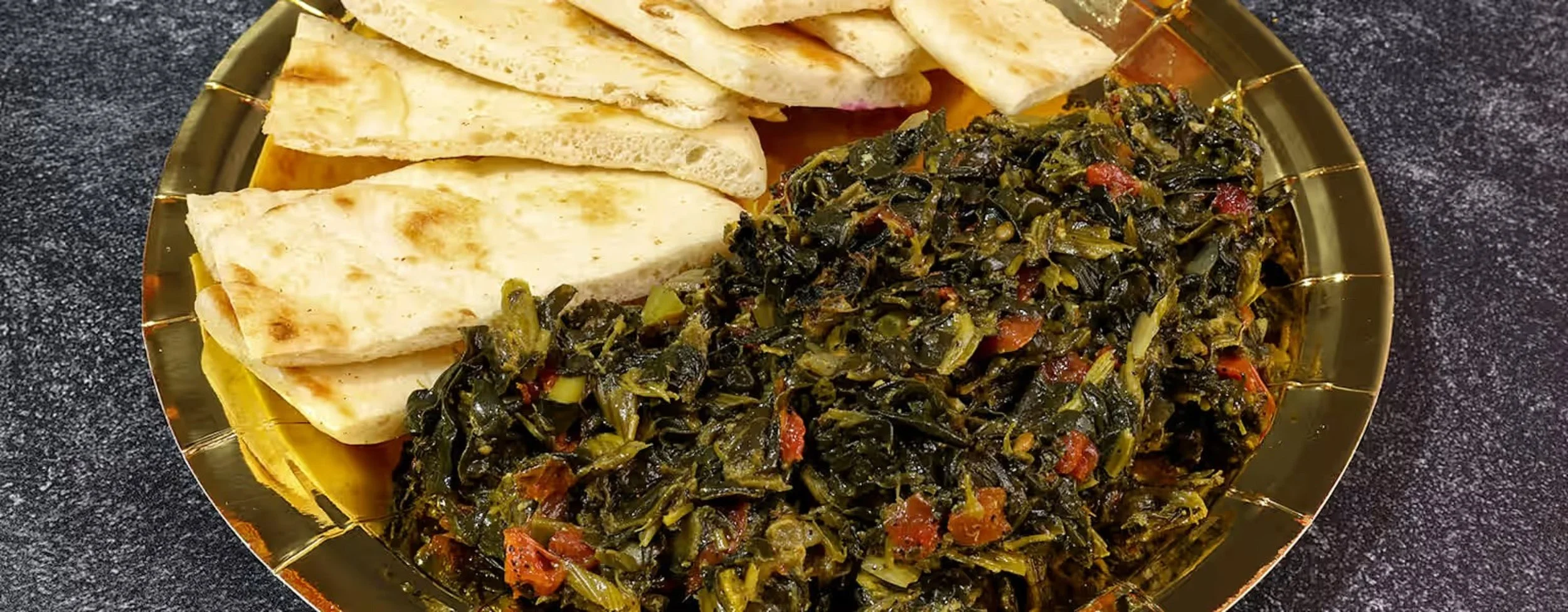A round golden tray with grilled pita bread and a serving of cooked leafy greens with vegetables. The tray is on a dark textured surface.