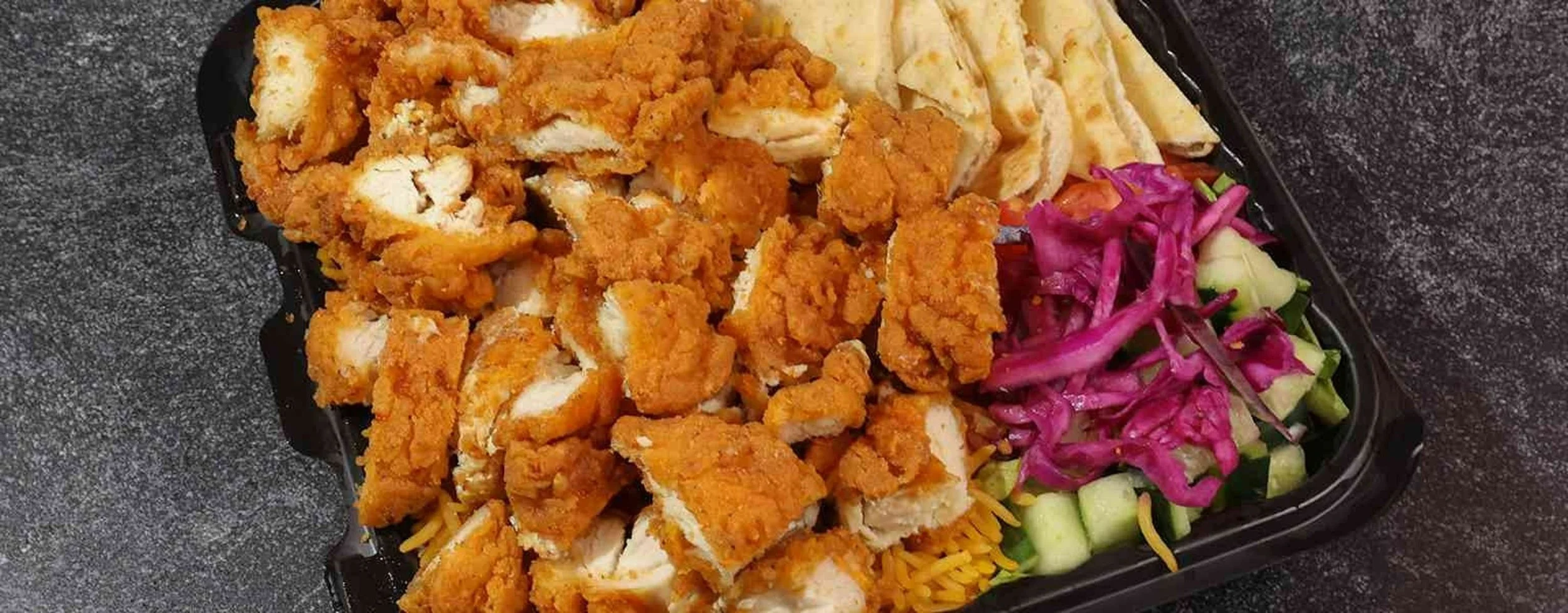 Fried chicken strips with shredded lettuce, purple cabbage, sliced cucumbers, and shredded cheese in a black plastic takeout container on a dark speckled surface.