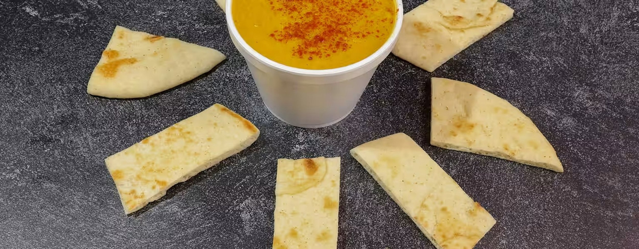 Six pieces of flatbread arranged in a circle around a cup of yellow soup with red spices, on a dark textured surface.
