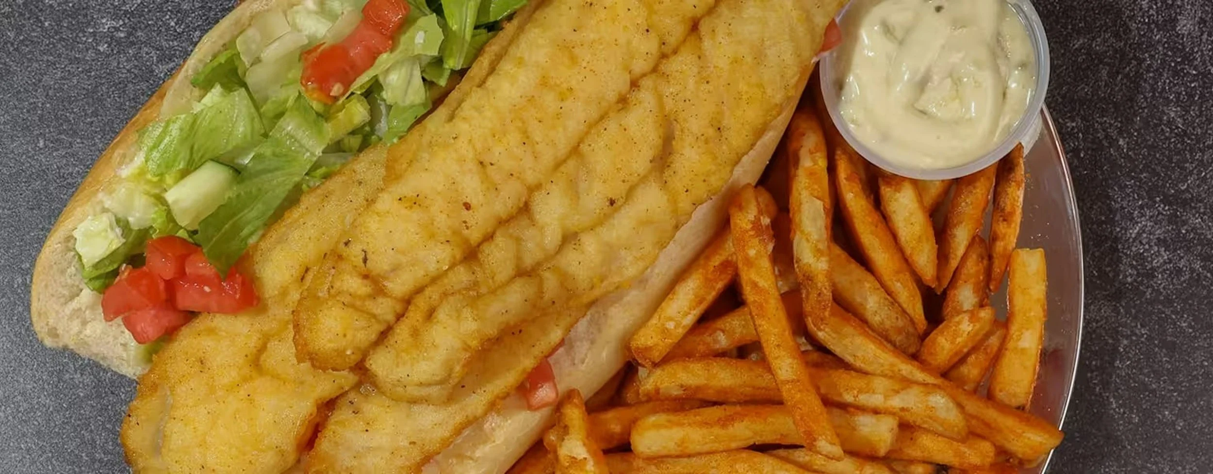 A plate of fried fish fillet sandwiches with French fries, a small cup of tartar sauce, shredded lettuce, chopped tomato, and pickles.