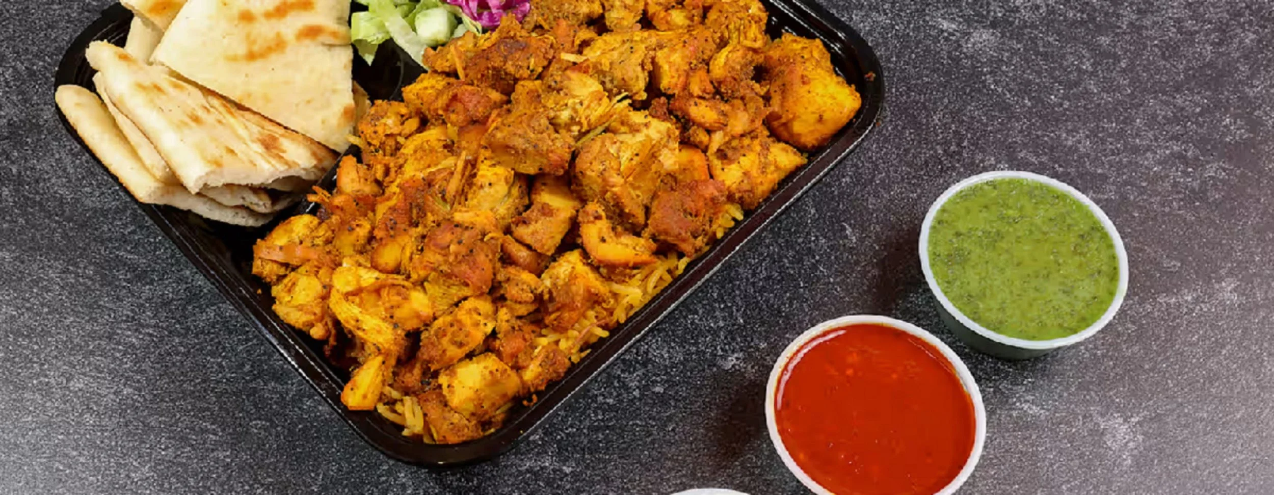 A black meal tray containing our Chicken Platter, with pita bread and a small portion of salad. Next to the tray are two cups of dipping sauces, one green and one red.