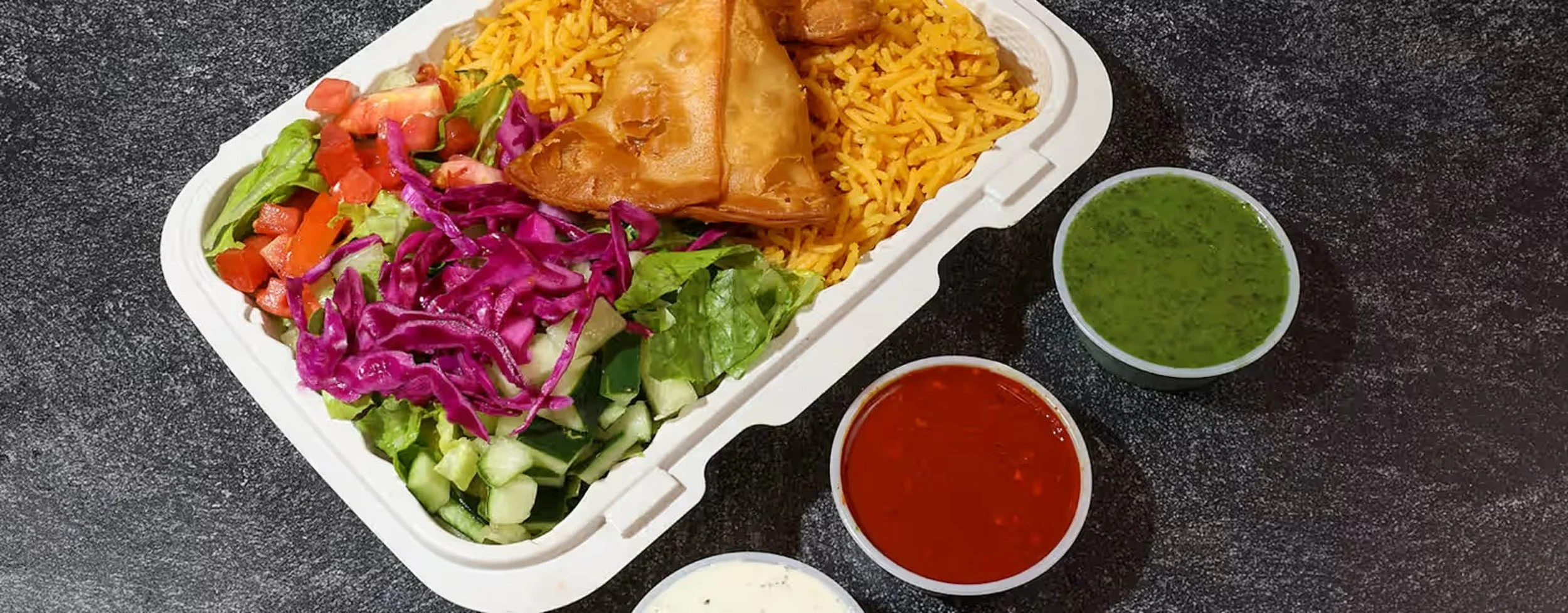 Takeout plate with yellow rice, samosas, fresh salad, and three containers of green, red, and white sauces on black textured surface.