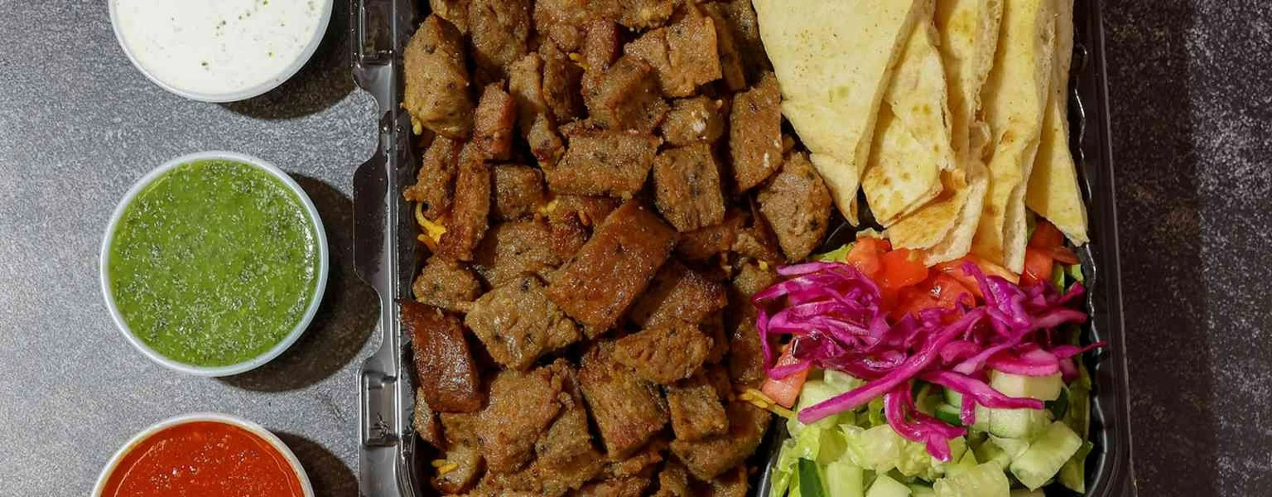 A takeout container with seasoned grilled meat, yellow flatbread, fresh salad with tomatoes and purple cabbage, and four small cups of sauces with green, white, and red condiments.