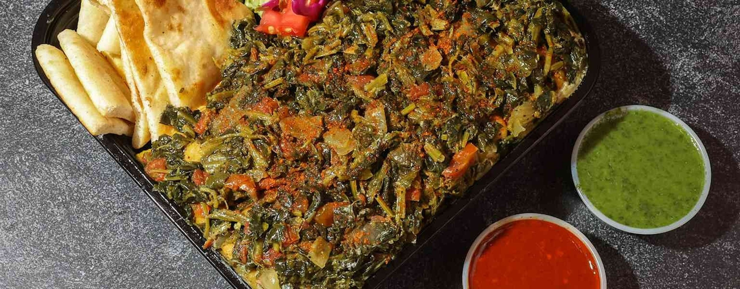 Plate of cooked greens with chopped carrots, potato pieces, French fries, and two small containers of green and red dipping sauces on a dark textured surface.