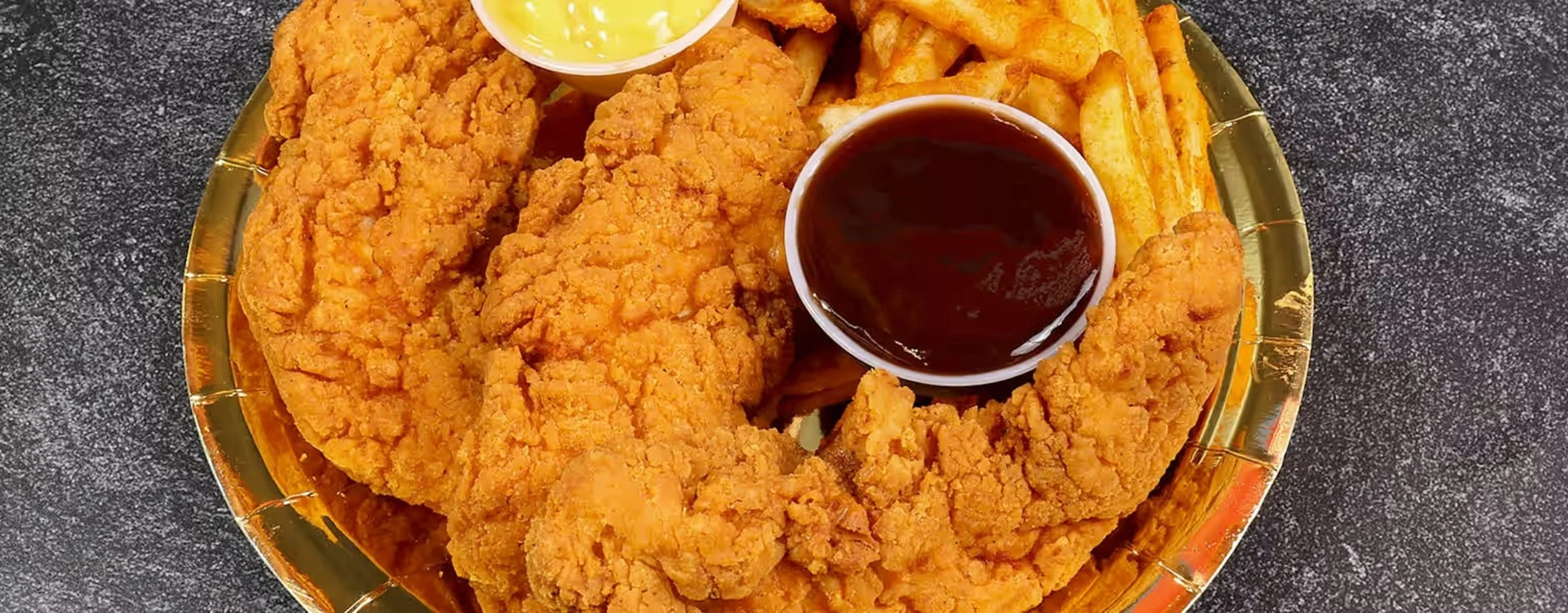 A plate of fried chicken tenders with French fries, dipping sauces including honey mustard and barbecue sauce on a dark surface.
