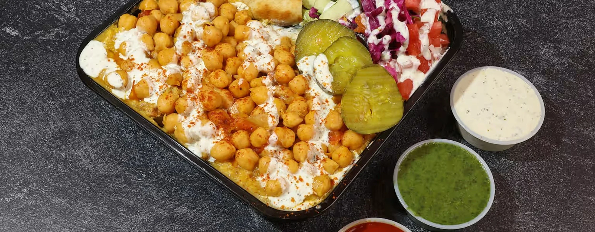 An overhead view of a baked dish with chickpeas, vegetables, and sauce, accompanied by three small bowls of sauce or dressing, on a dark textured surface.