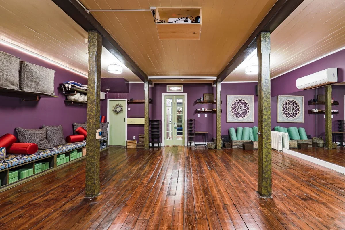 Empty yoga studio with wooden floors, purple walls, and mirrored wall on the right. There are various stuffed pillows and cushions on benches along the walls, and decorative wall art. Pillar posts support the ceiling, with some greenery. A door at th