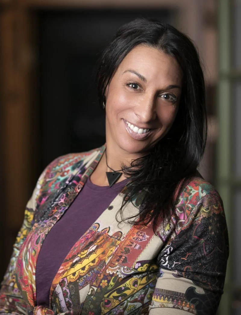A woman with long dark hair smiling, wearing a colorful, patterned blazer over a purple top, standing indoors near a window.