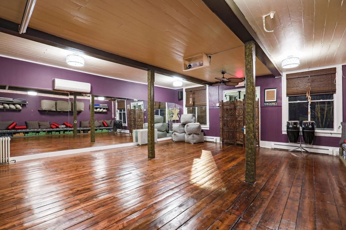 A spacious dance or yoga studio with wooden floors, purple walls, large mirrors, and fitness equipment, illuminated by ceiling lights.