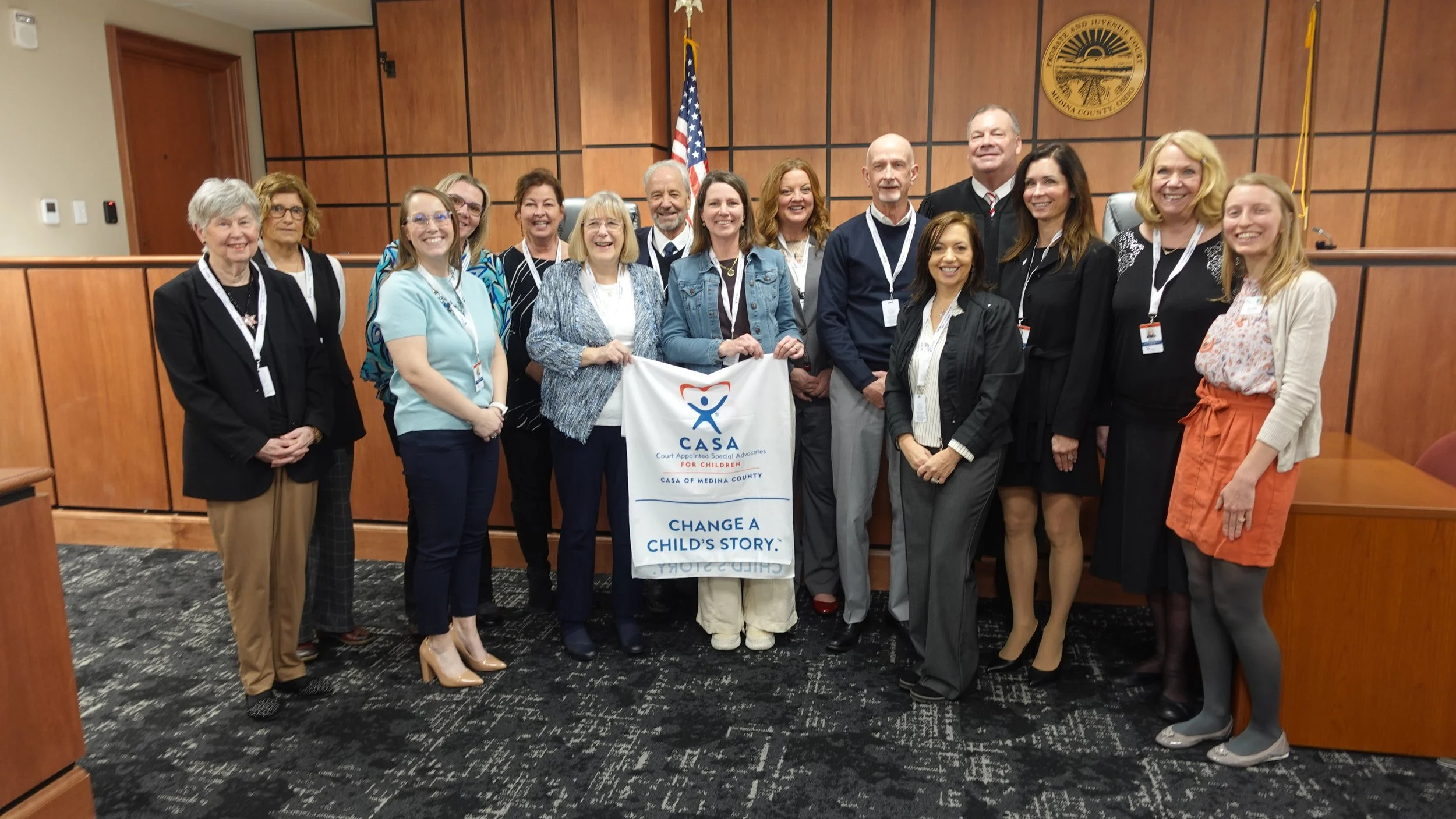 Group of new CASA volunteers standing in a courtroom, holding a CASA banner that reads 'Court Appointed Special Advocates for Children, CASA of Medina County, Change a Child's Story.'