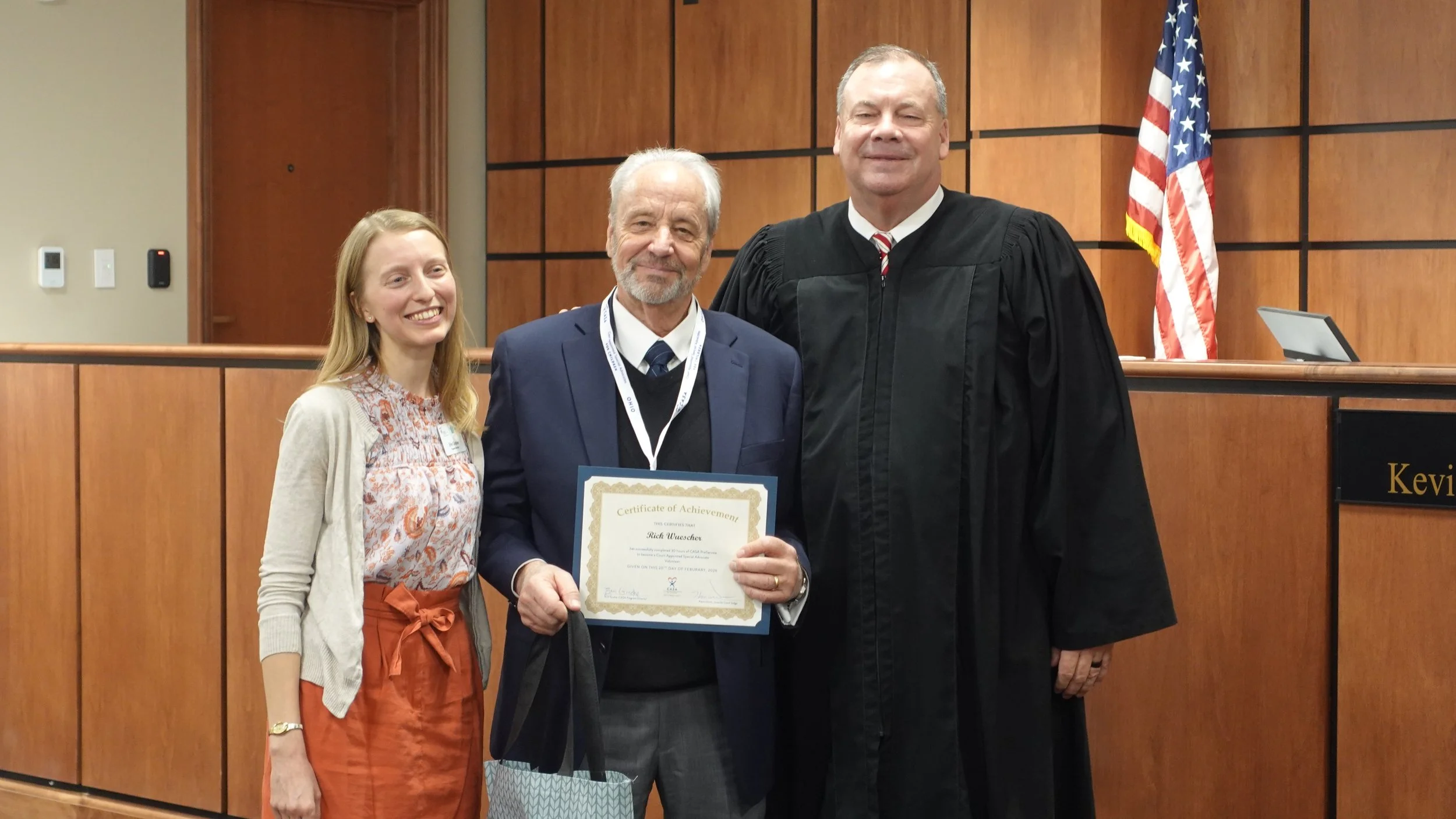 Three people standing in a courtroom during a volunteer swearing-in ceremony. A woman stands on the left, a man who just became a CASA volunteer in the middle, and the judge on the right.