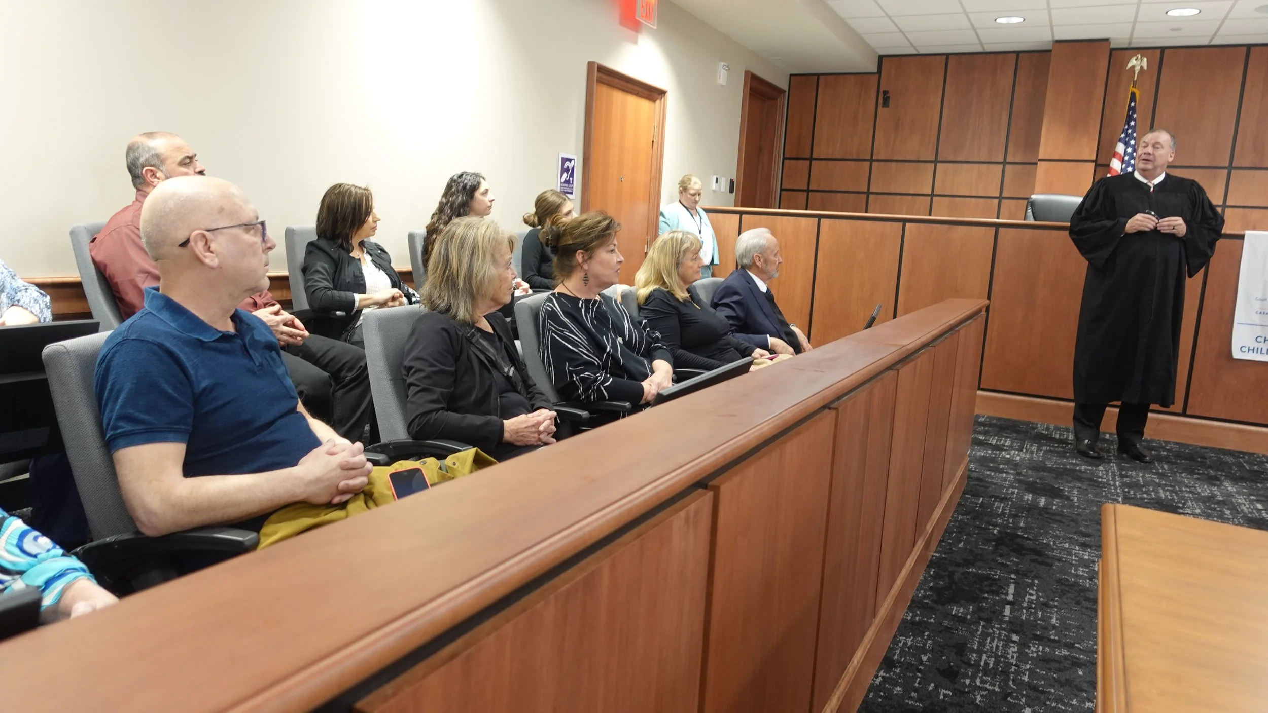 Courtroom with a judge in a black robe speaking to a seated audience of adults, including men and women, in a wood-paneled room with an American flag during a volunteer swearing-in ceremony.