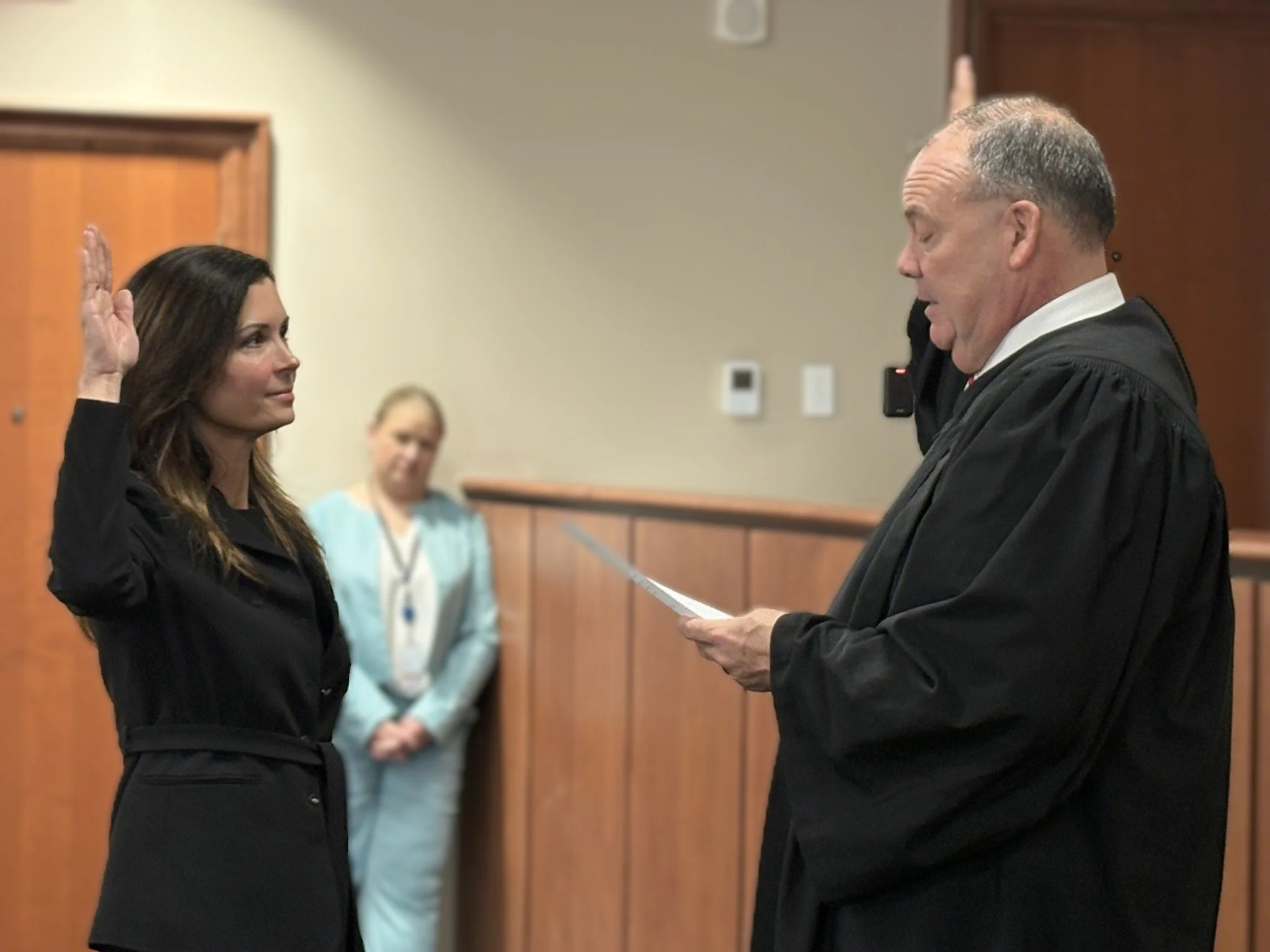 A woman in a black dress is raising her right hand during a court proceeding while an older male judge in robes reads from a document.