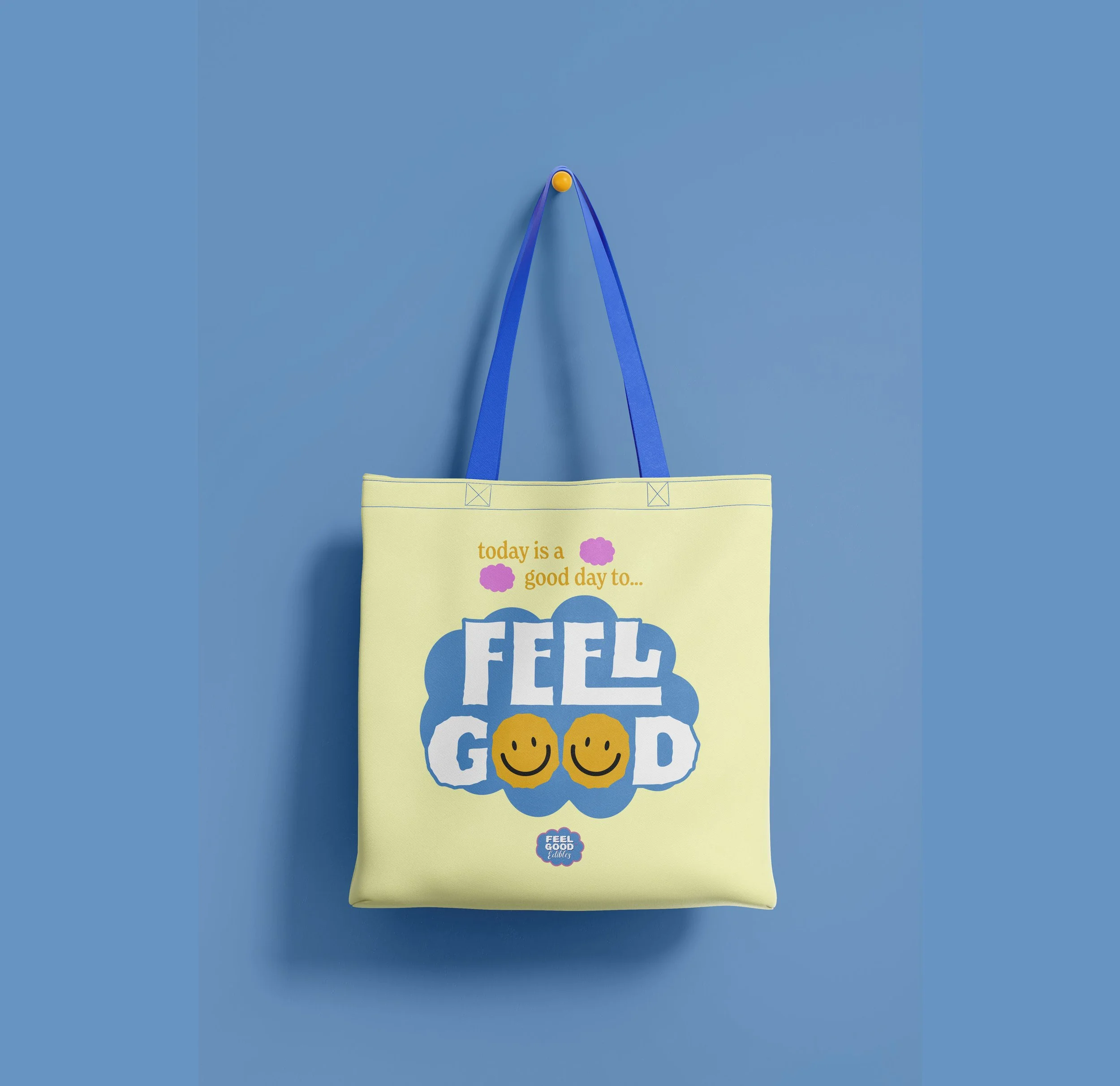 Yellow tote bag hanging on a blue wall with the message 'today is a good day to... FEEL GOOD' and two smiley faces