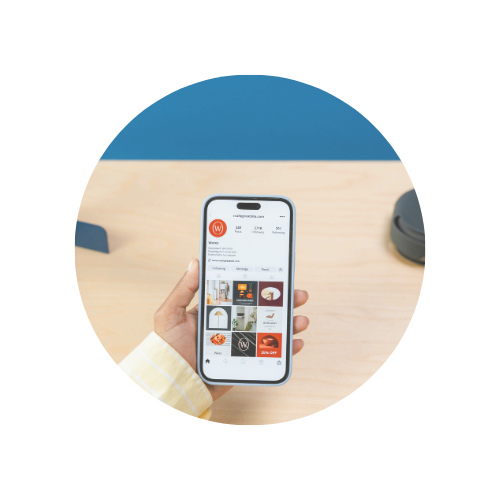 A person holding a smartphone displaying an app with a red circular logo and food images, on a wooden table with a black object nearby.