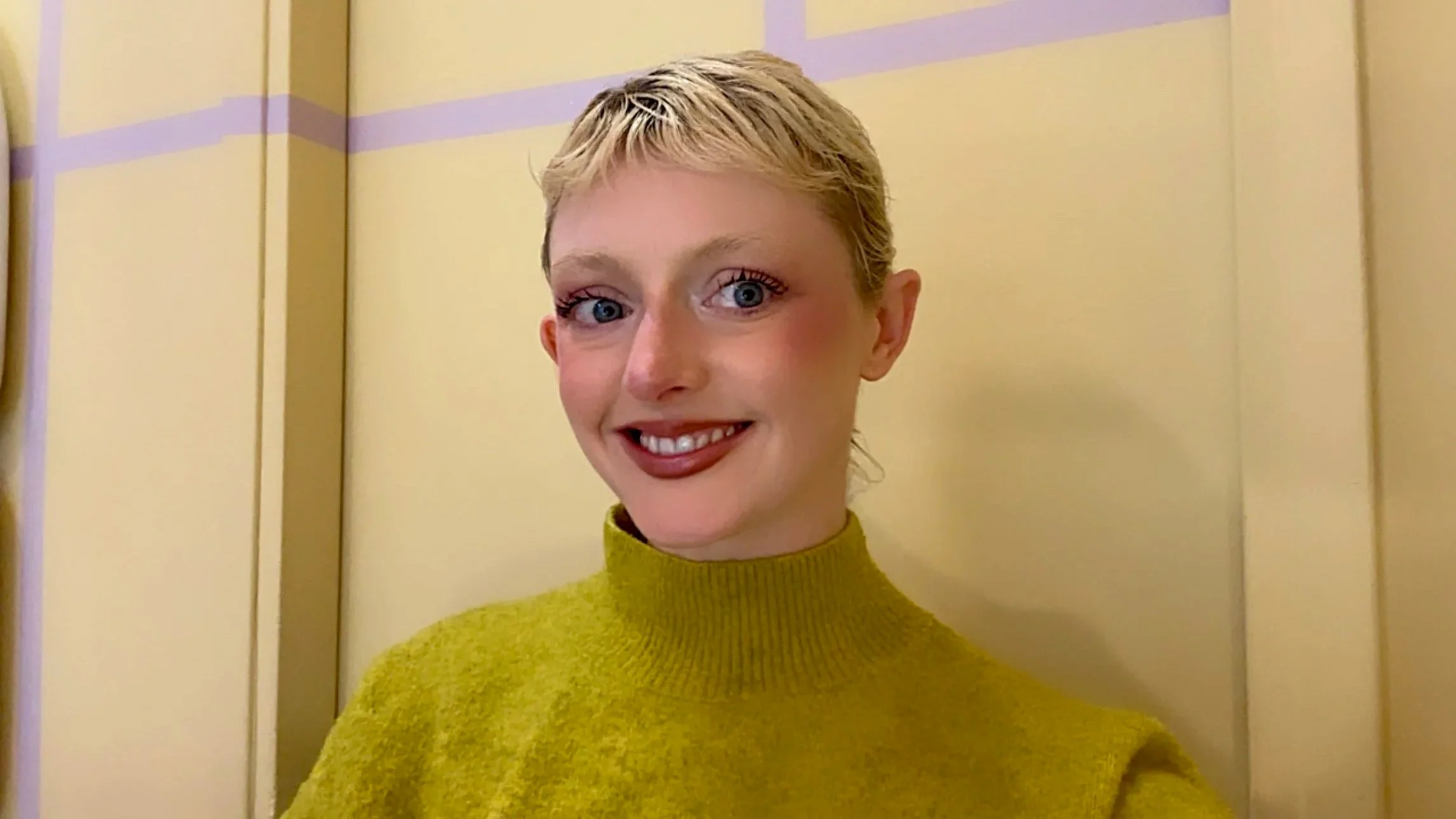 A young woman with short blonde hair, blue eyes, and a big smile, wearing a yellow turtleneck sweater, standing against a light-colored wall with purple horizontal stripes.