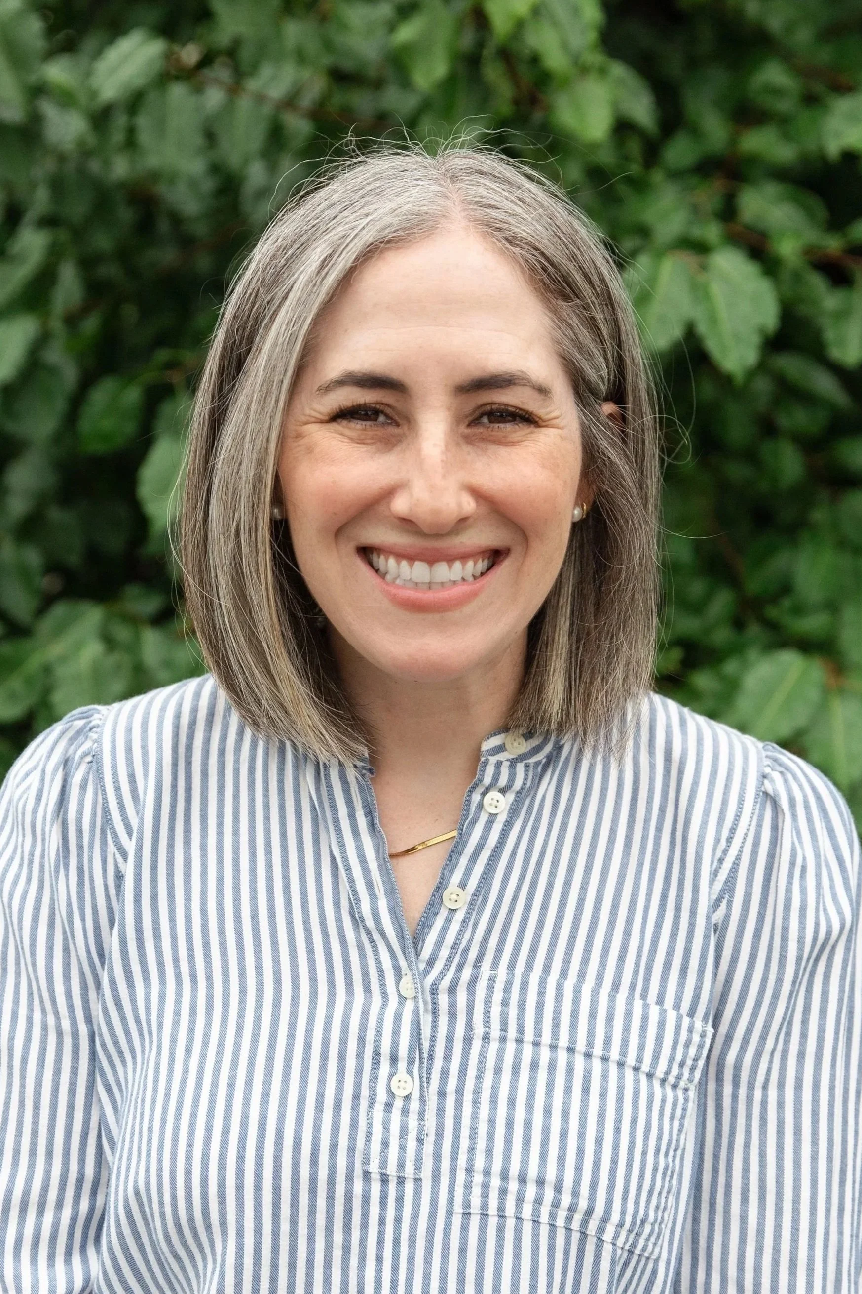 A smiling woman with shoulder-length gray hair, wearing a striped button-up shirt, standing outdoors with green foliage in the background.