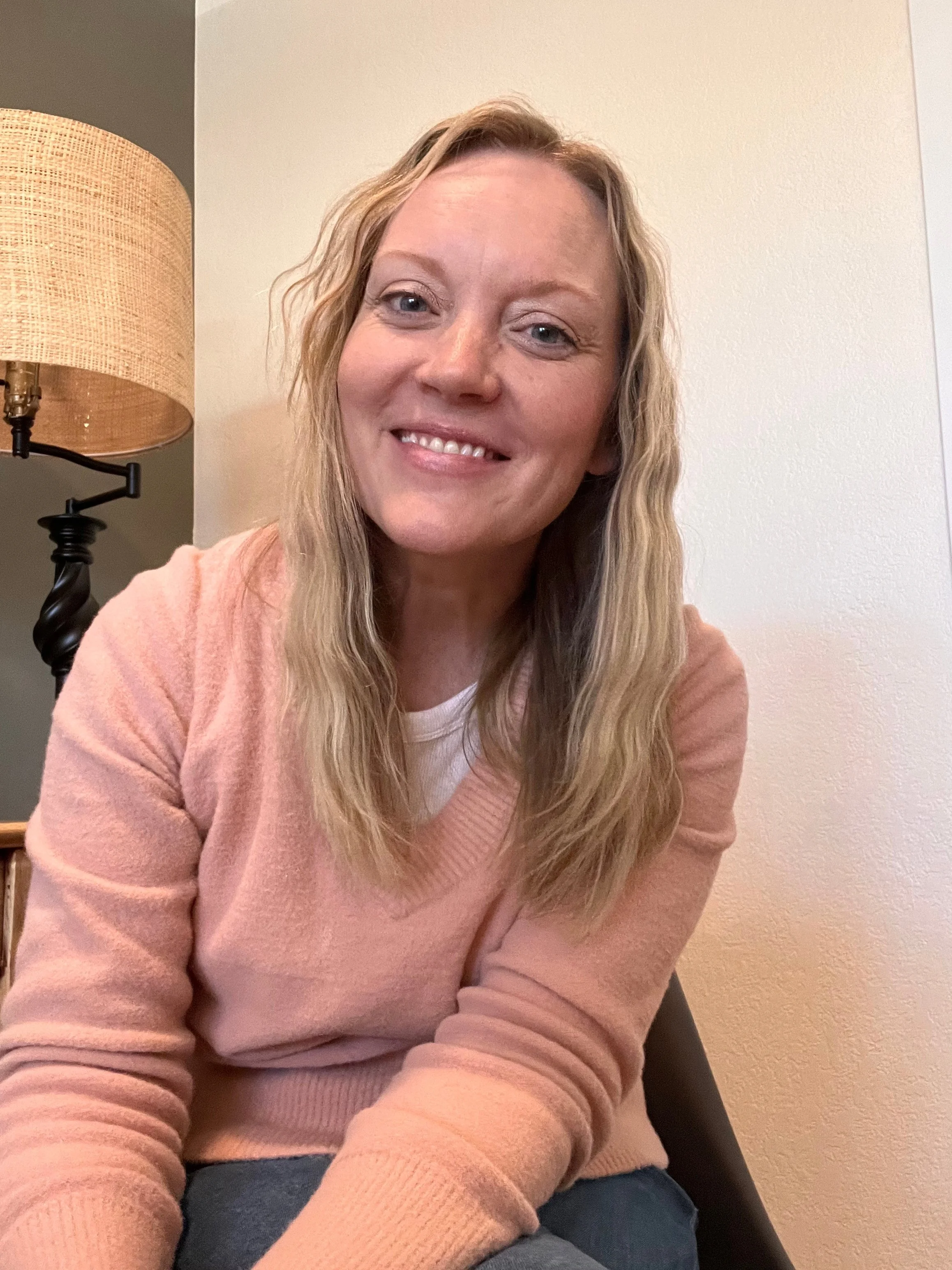 Smiling woman with long blonde hair wearing a pink sweater, sitting indoors near a beige wall with a lamp in the background.