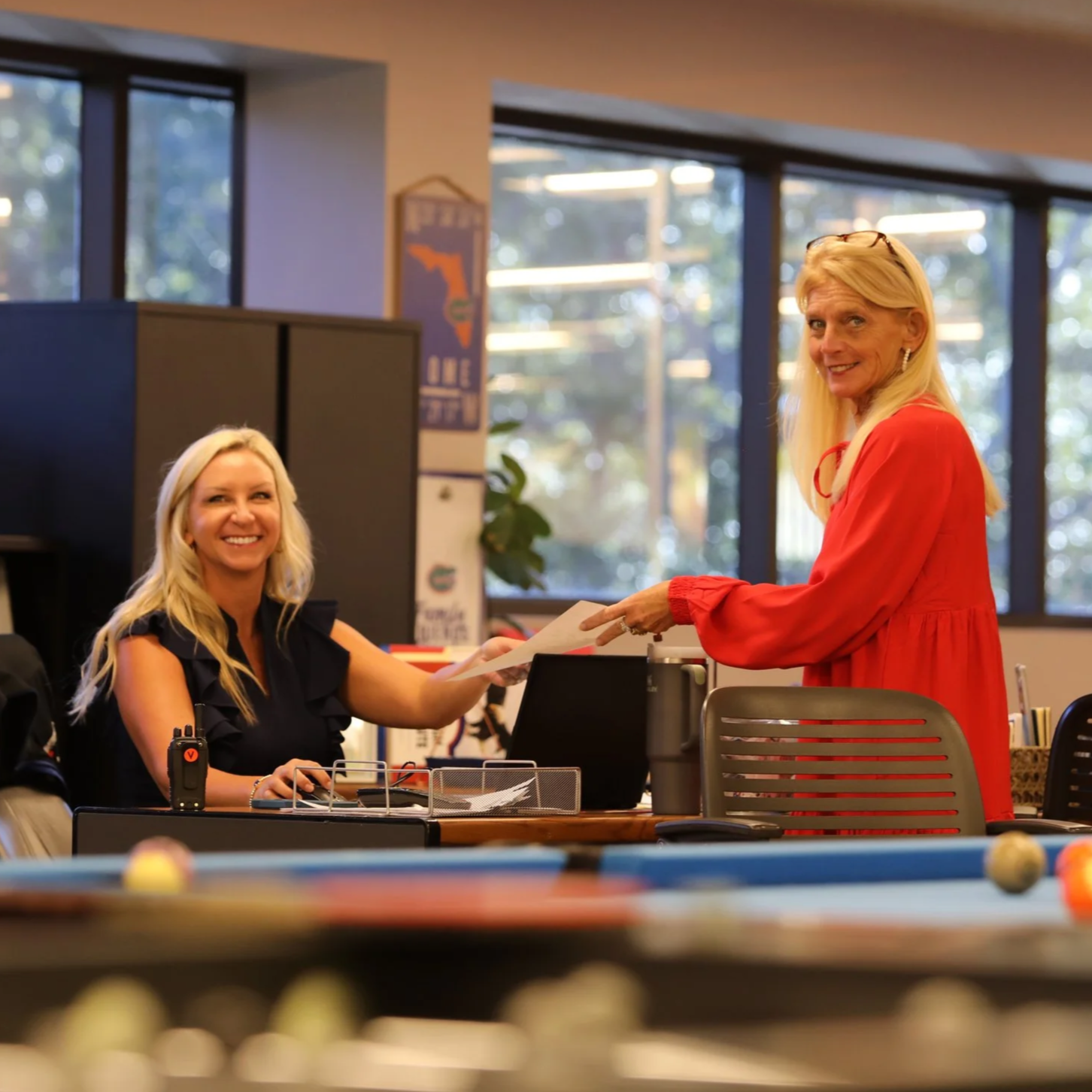 Two women in an office or classroom setting, one seated at a desk and the other standing, exchanging a document. The woman standing wears a red dress, and the woman sitting wears a black sleeveless top, both smiling.